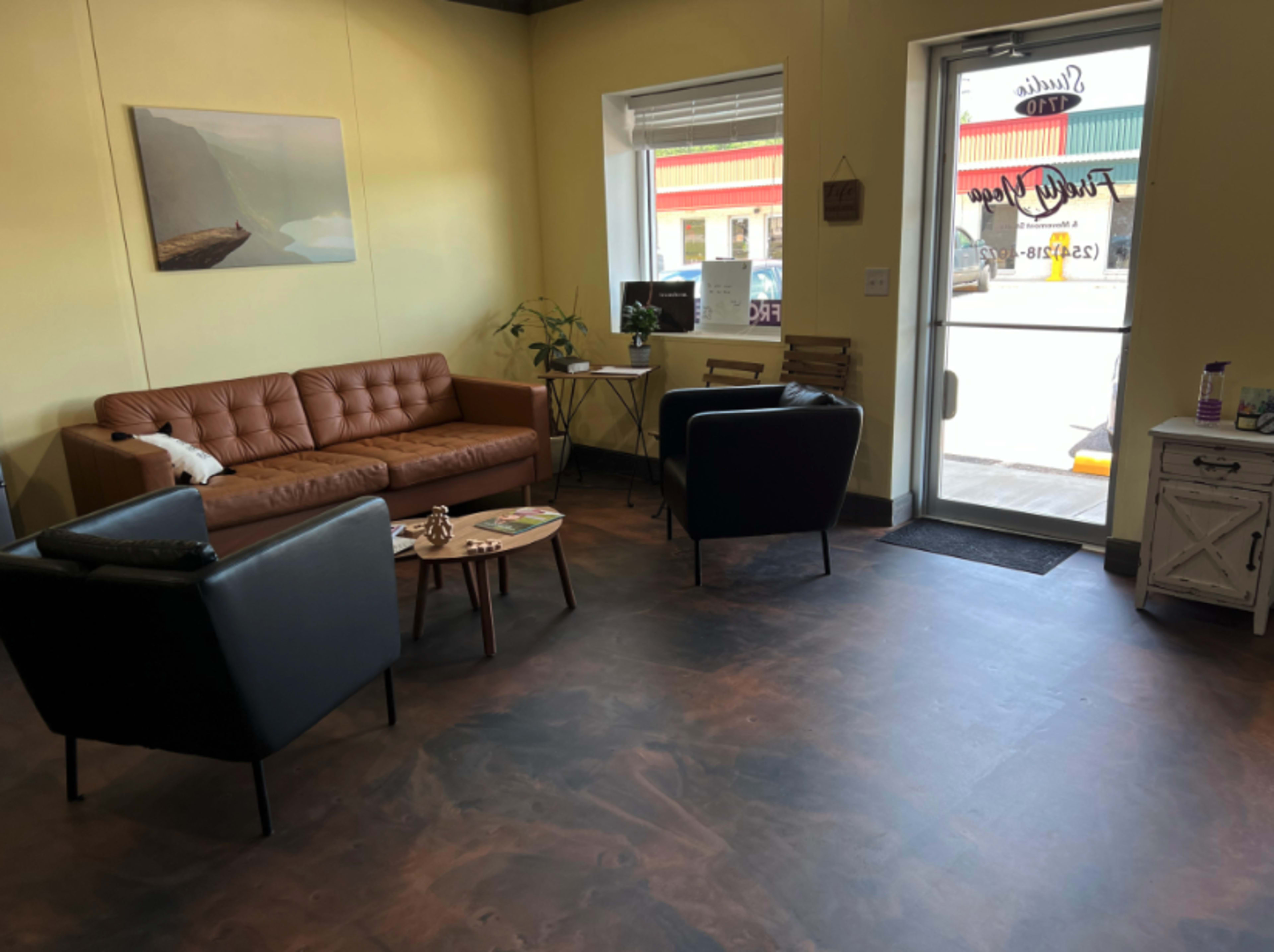 A cozy waiting area with a brown leather couch, two black chairs, a small table, and a door leading outside.