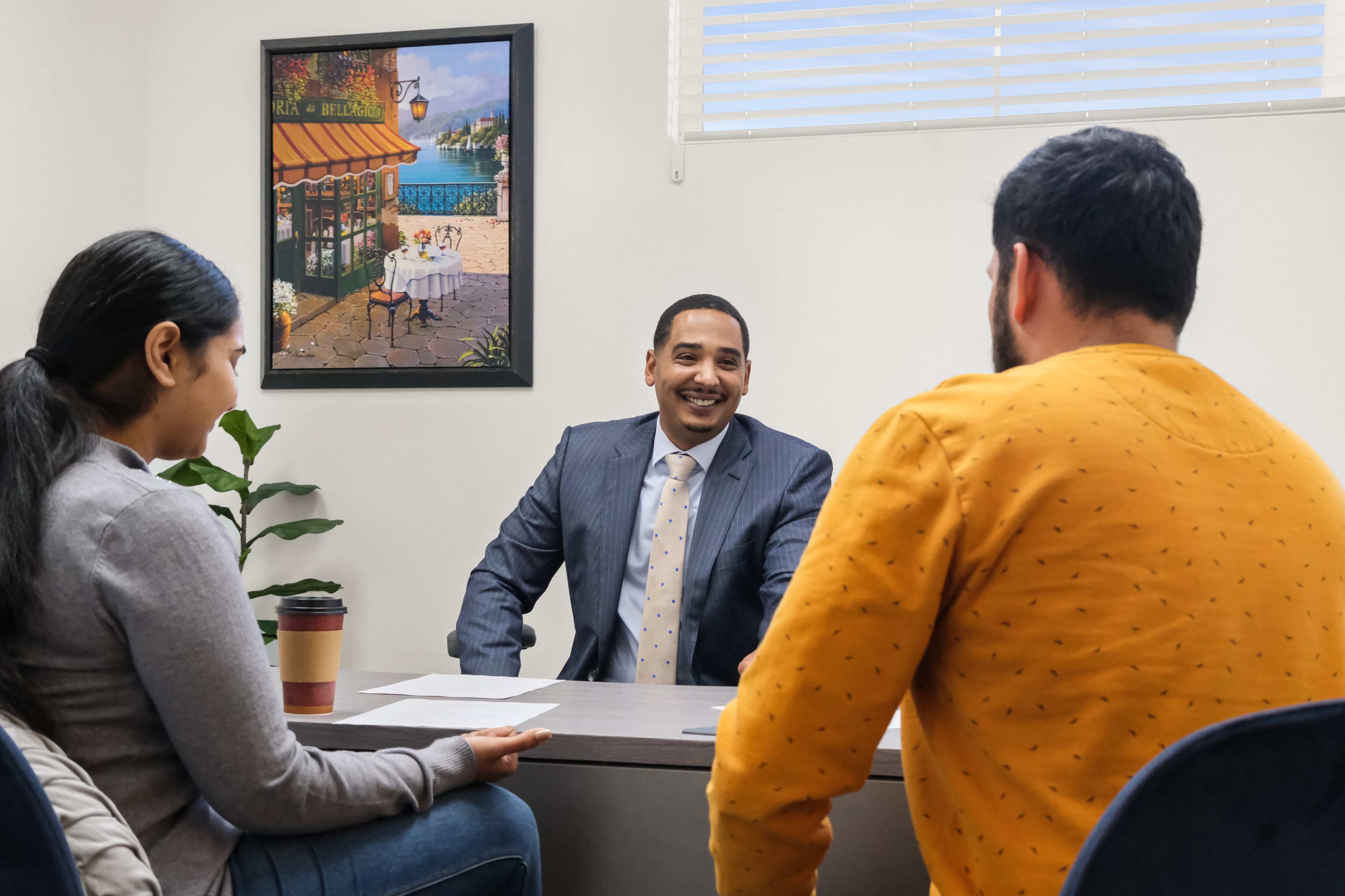 A man in a suit sits at a table facing two people, one wearing a gray sweater and the other a mustard-colored sweater, in a well-lit office with a plant and a colorful painting on the wall.