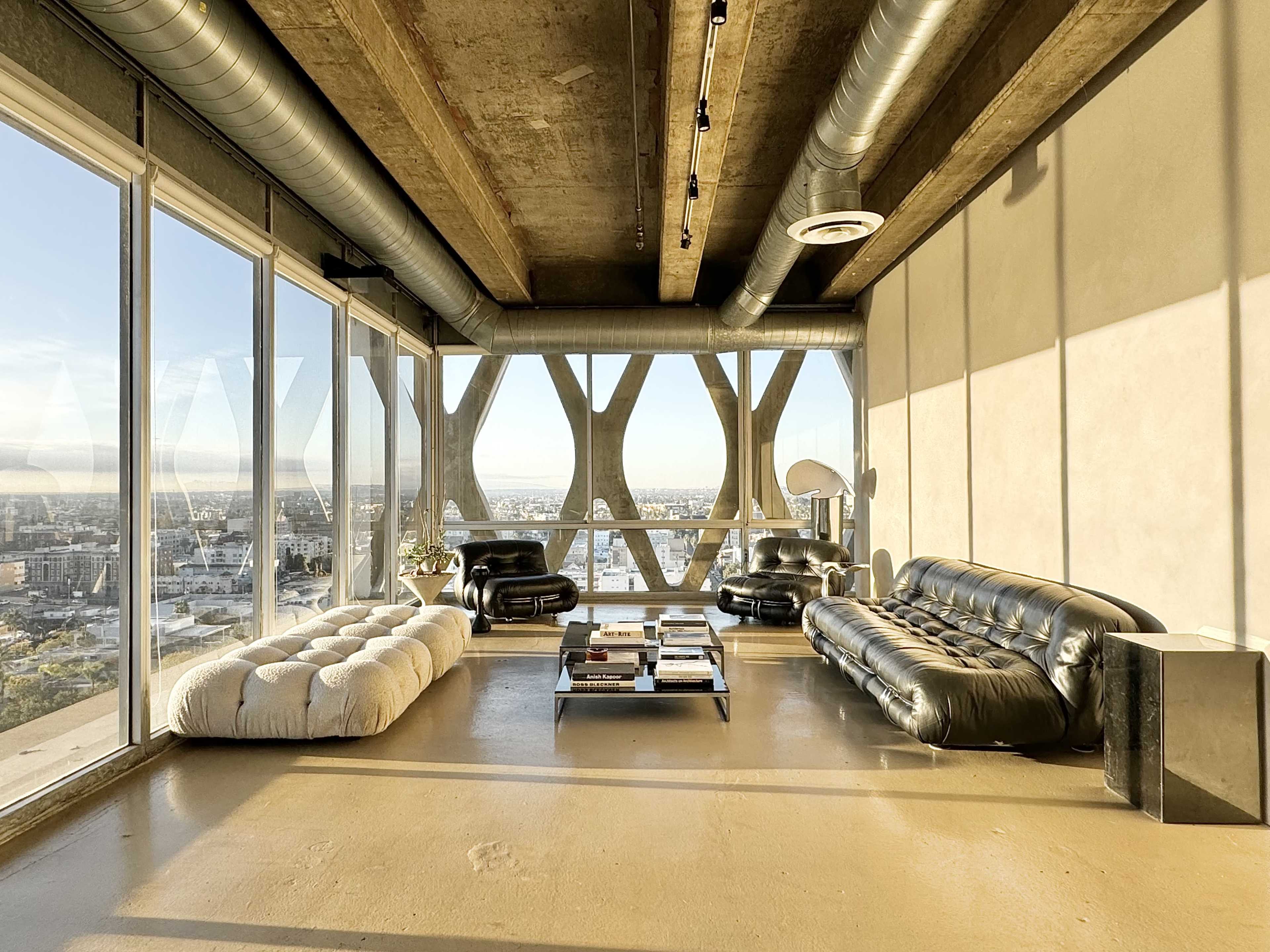 A modern living room features two black leather sofas and a coffee table, with large windows showcasing a city view against a backdrop of exposed concrete and metal beams.