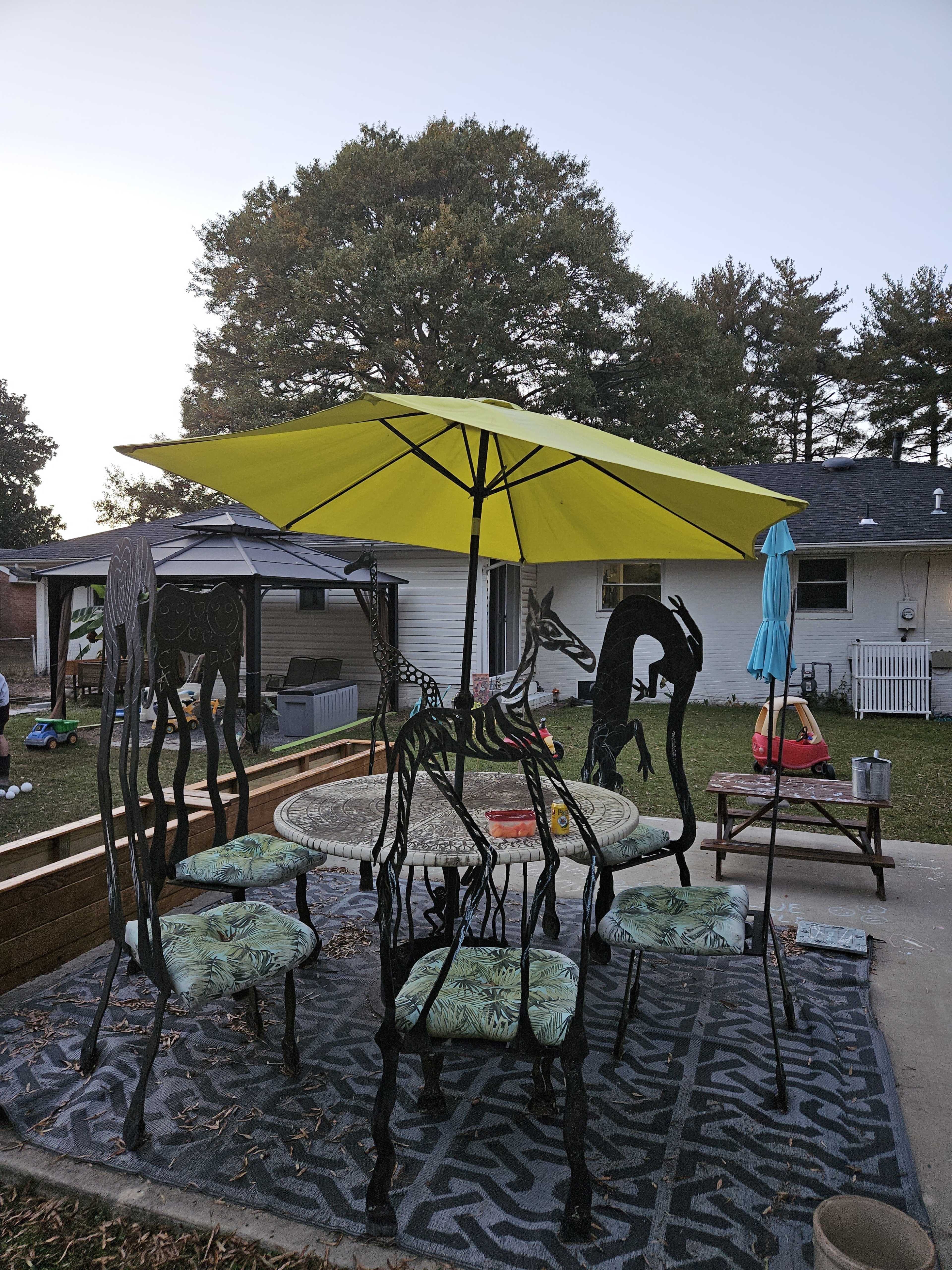 A black metal patio set with a round table and colorful patterned chairs is shaded by a yellow umbrella in a backyard setting.