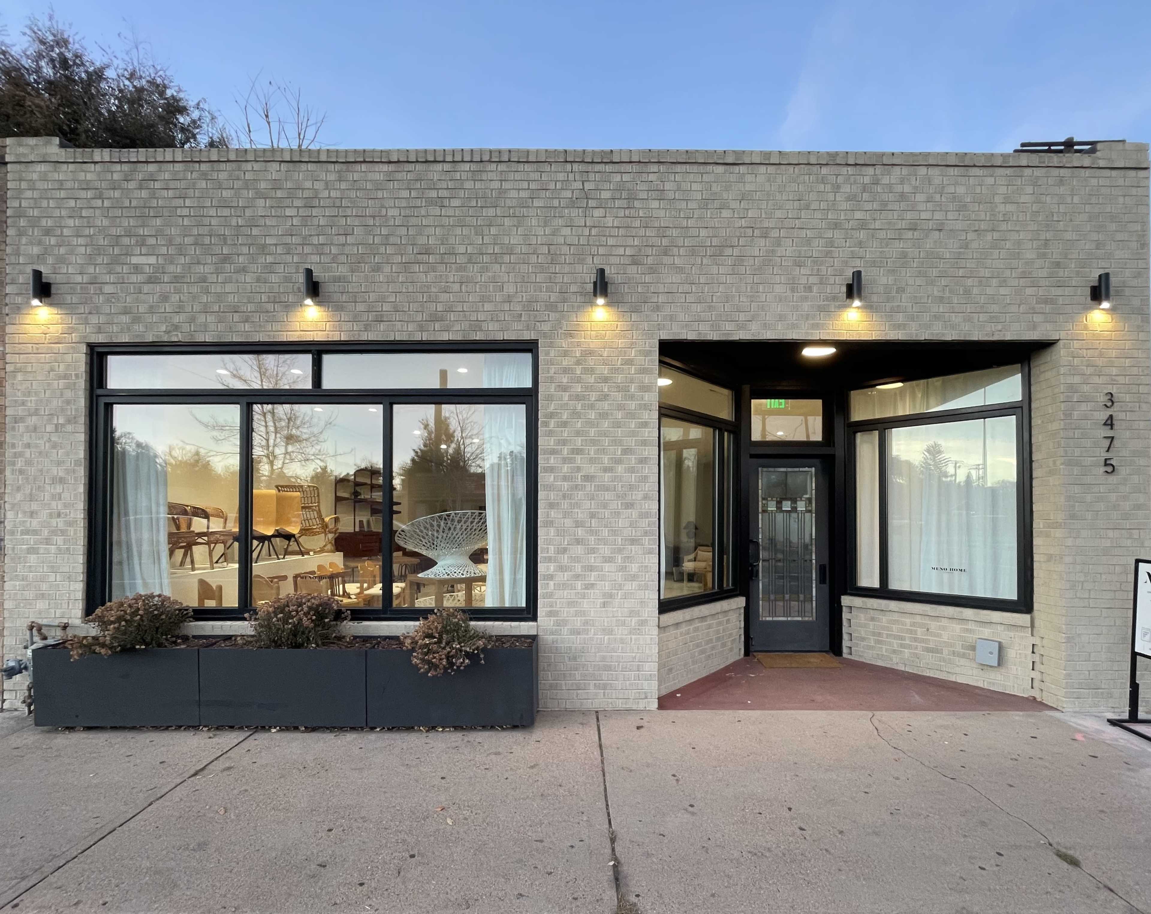 A one-story building featuring large windows, a glass front door, and a minimalist exterior design with potted plants.