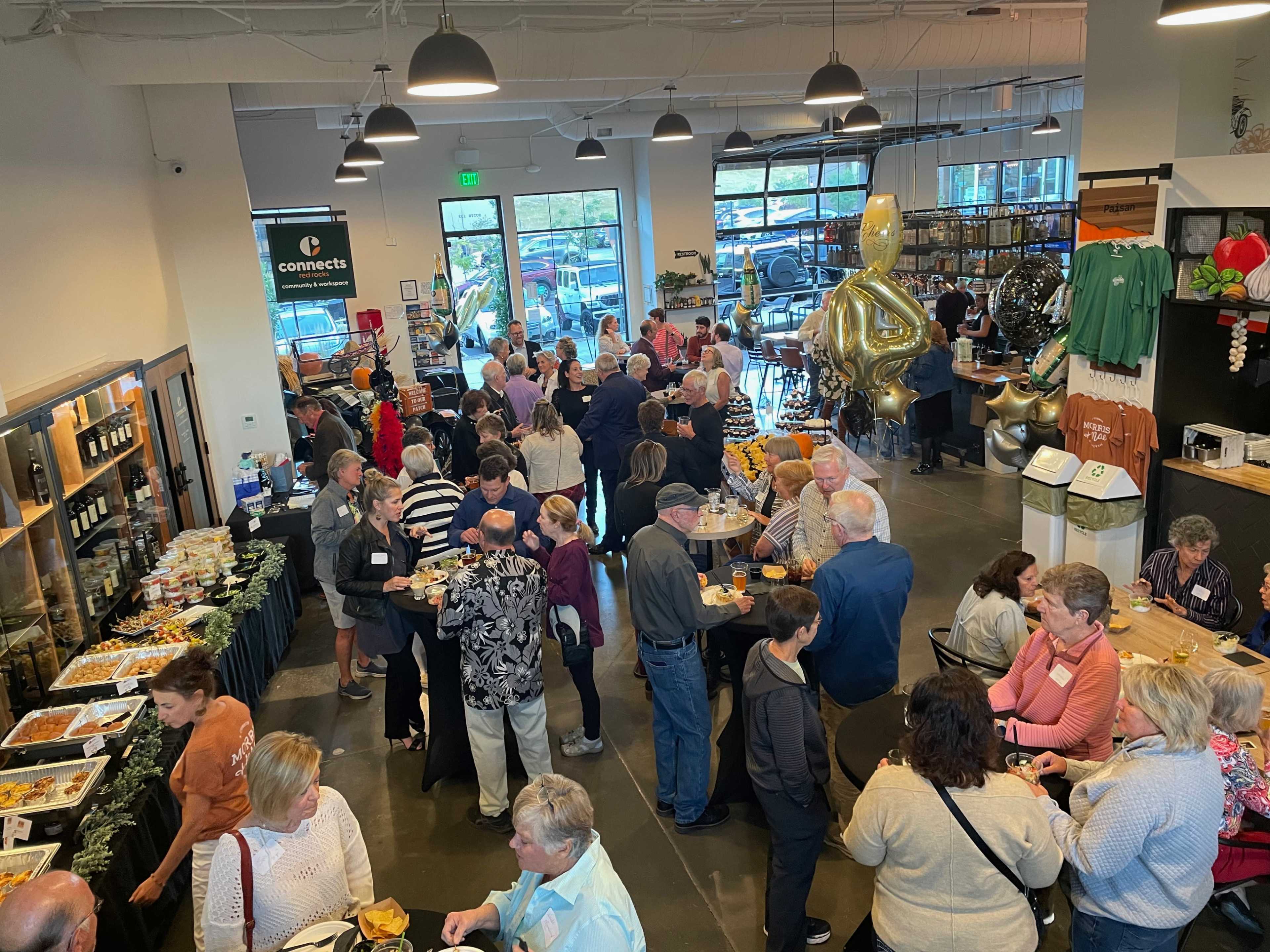 A large crowd gathers in a spacious indoor venue filled with tables, food stations, and decorations, indicating an event or gathering.