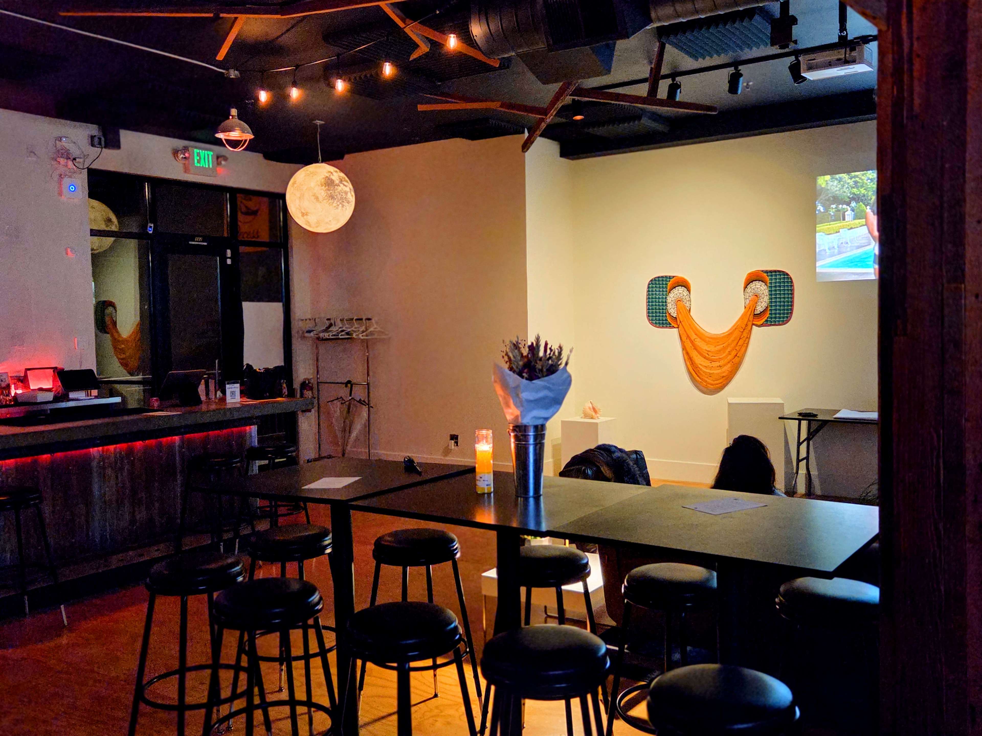 The image shows a modern, dimly lit bar area with high tables and stools, featuring a decorative wall with art and a projection screen.