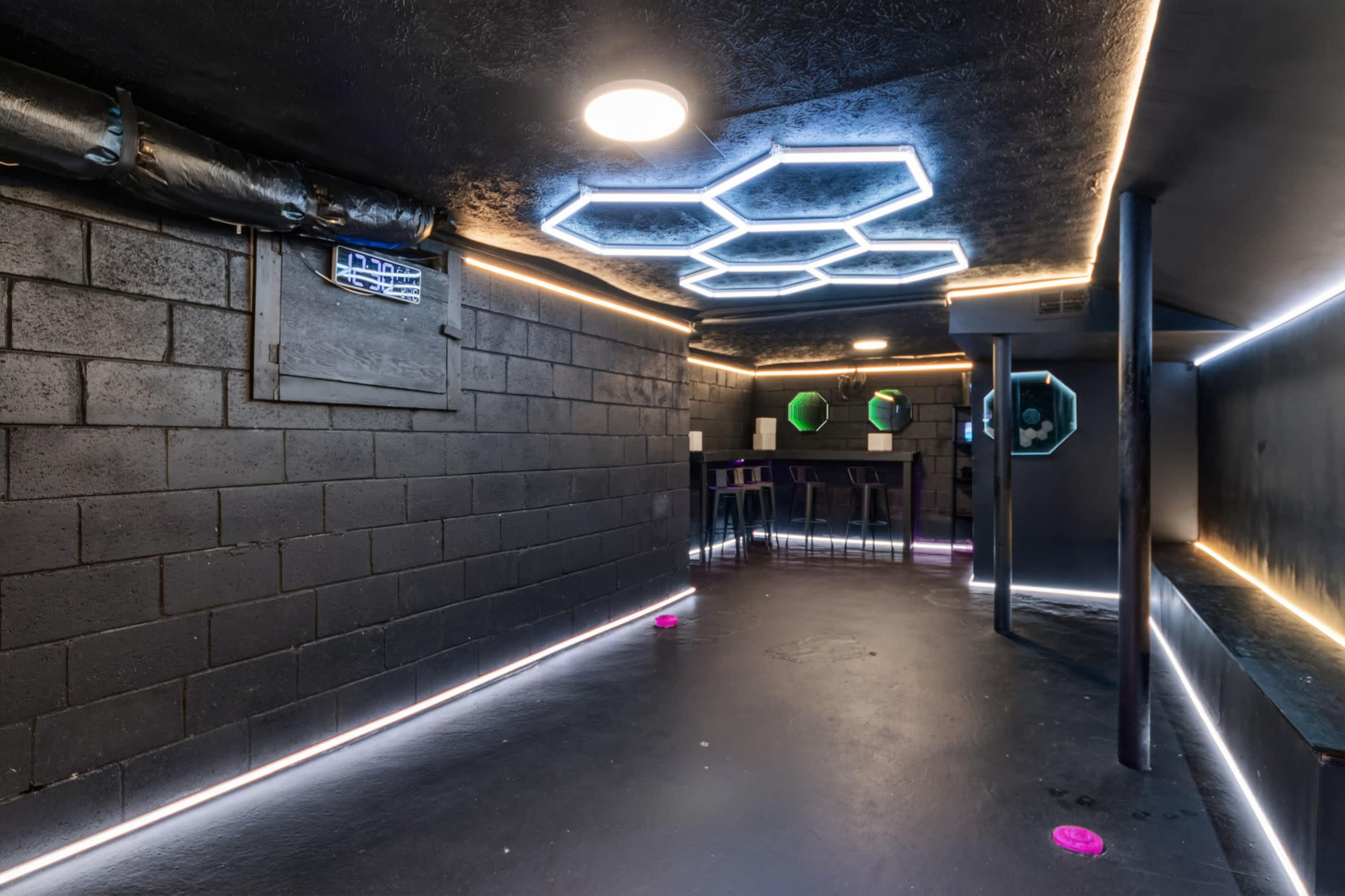 The image shows a dimly lit basement space with black walls, geometric LED lighting, and a bar-like table along one side.