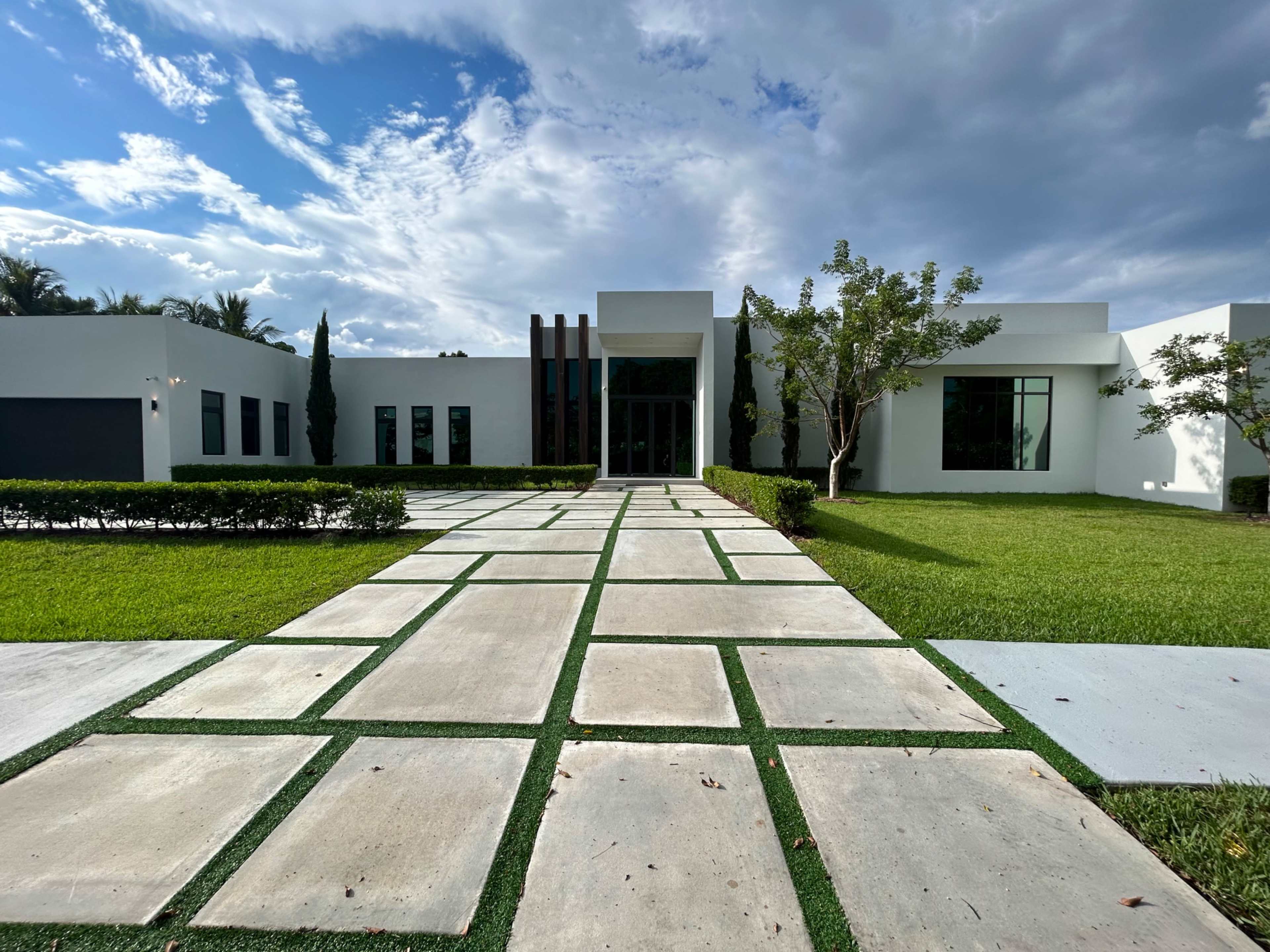 A modern house with a paved pathway and neatly trimmed landscaping leads to the entrance, set against a backdrop of cloudy skies.