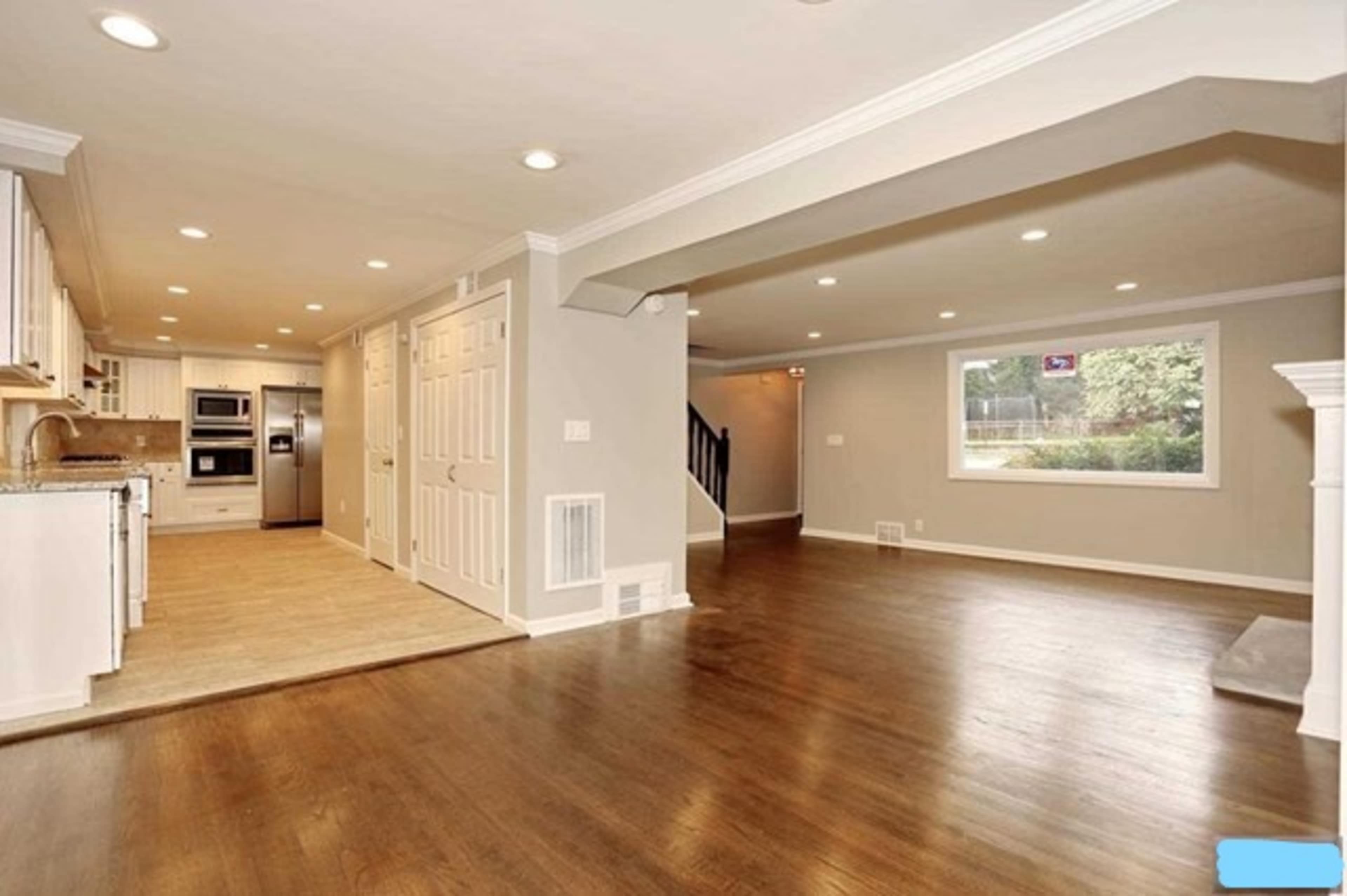 A spacious, open-concept living area with hardwood floors, a large window, and a modern kitchen featuring white cabinetry and stainless steel appliances.