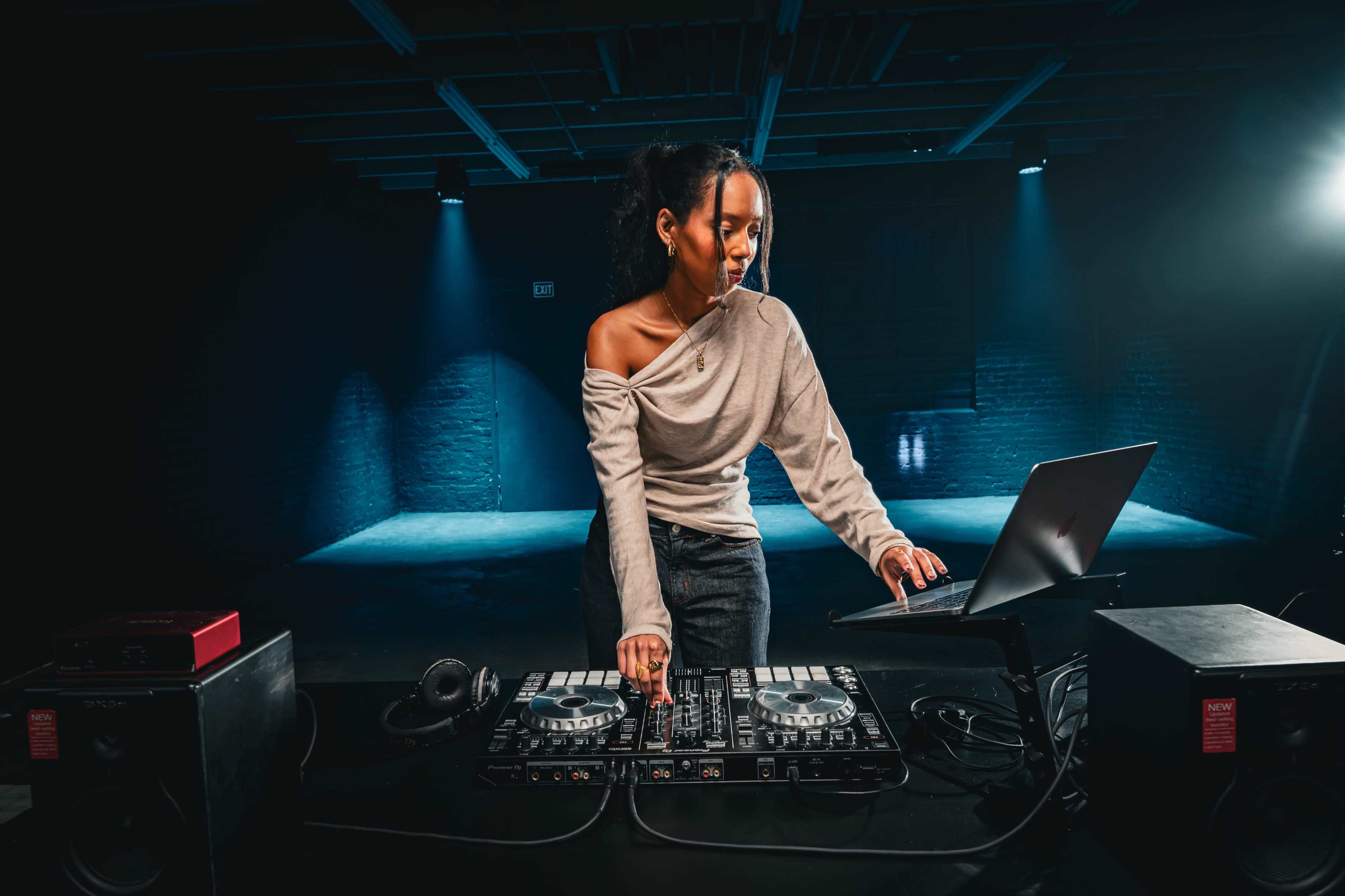 A DJ stands at a mixer with a laptop in a dimly lit studio, surrounded by speakers and stage lights.
