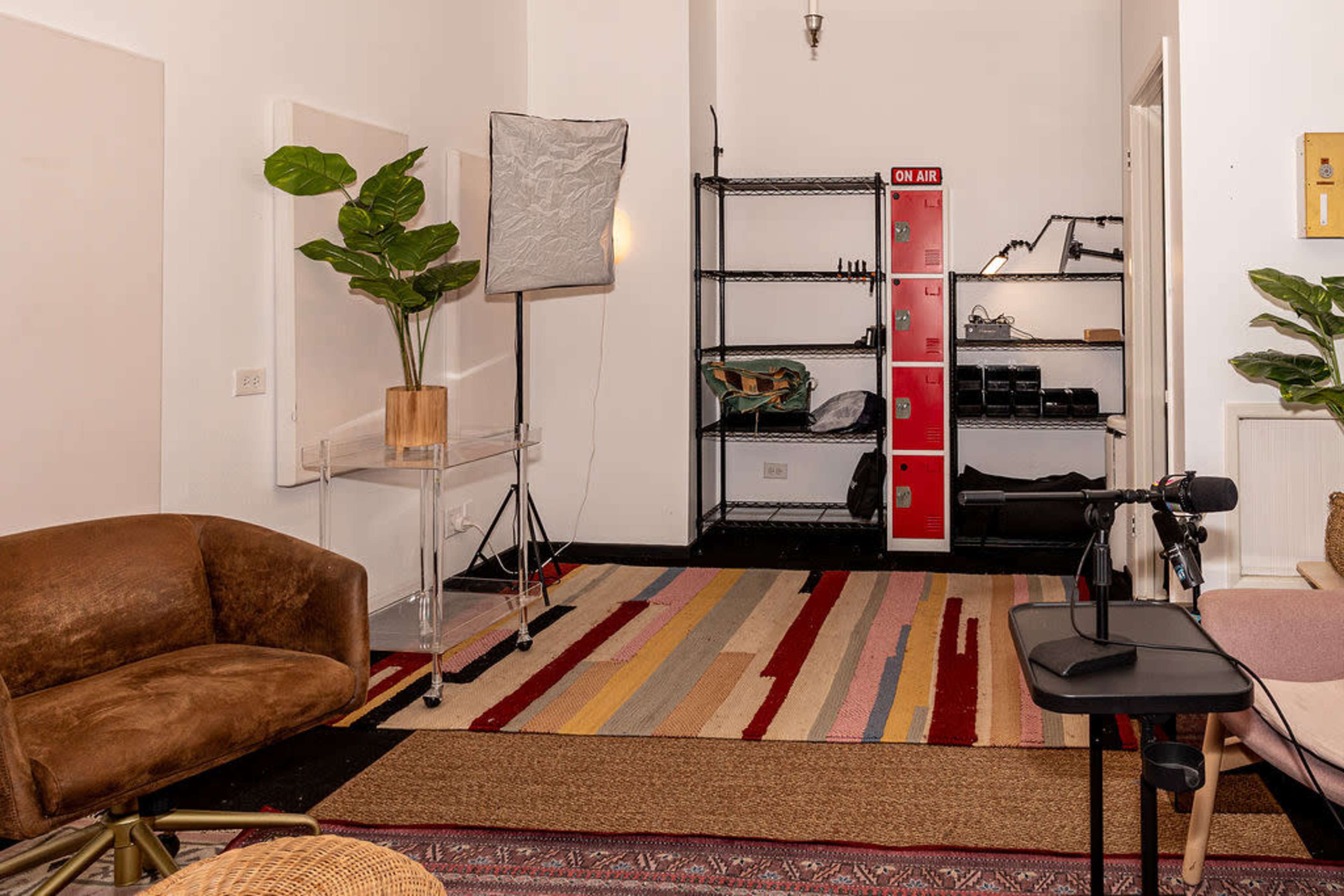 The image shows a small studio space featuring a brown upholstered chair, a clear table, and a microphone setup, alongside shelves of equipment and a potted plant.
