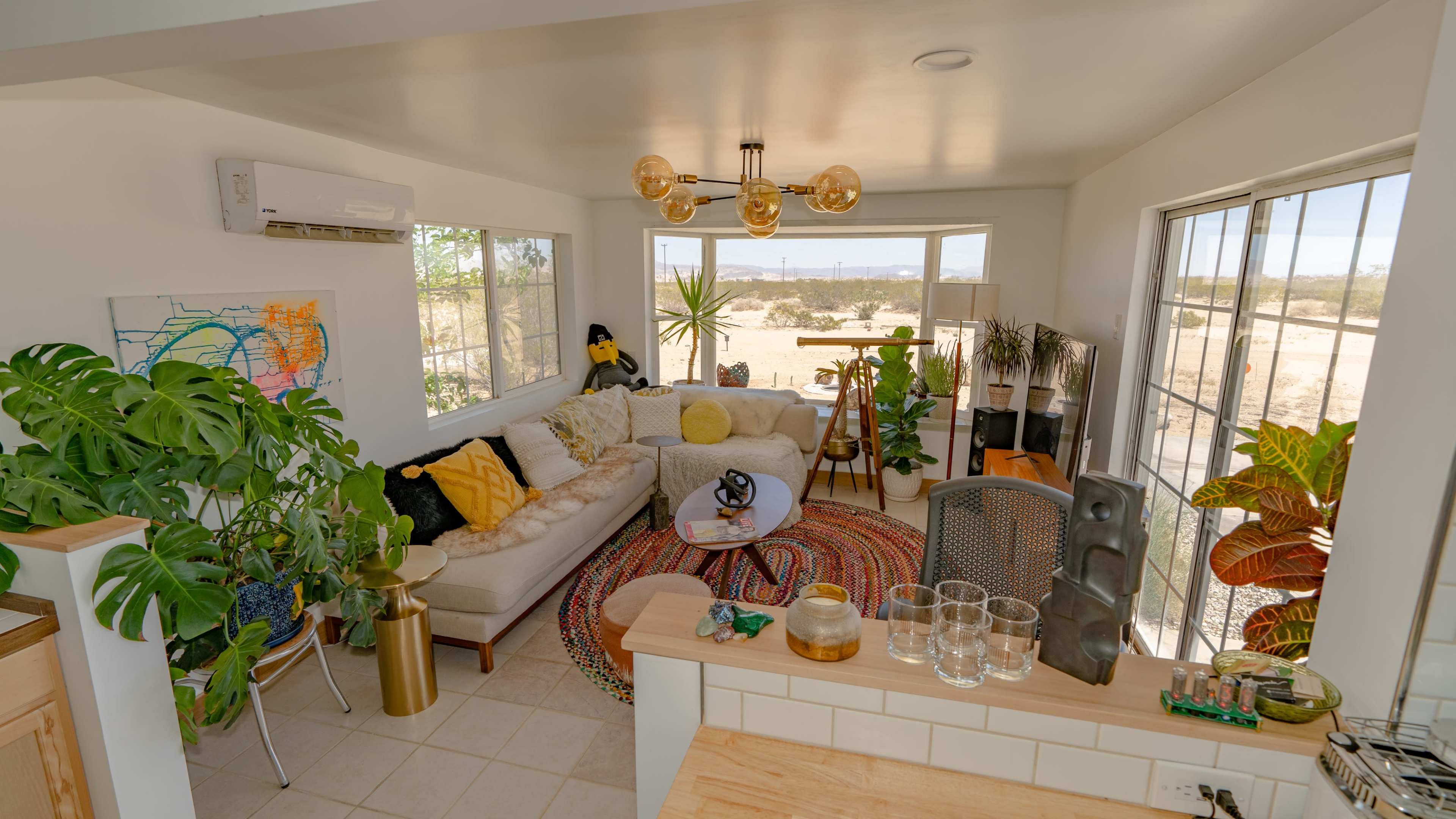 The image shows a modern living room with a large sectional sofa, a rug, plants, and windows overlooking a desert landscape.