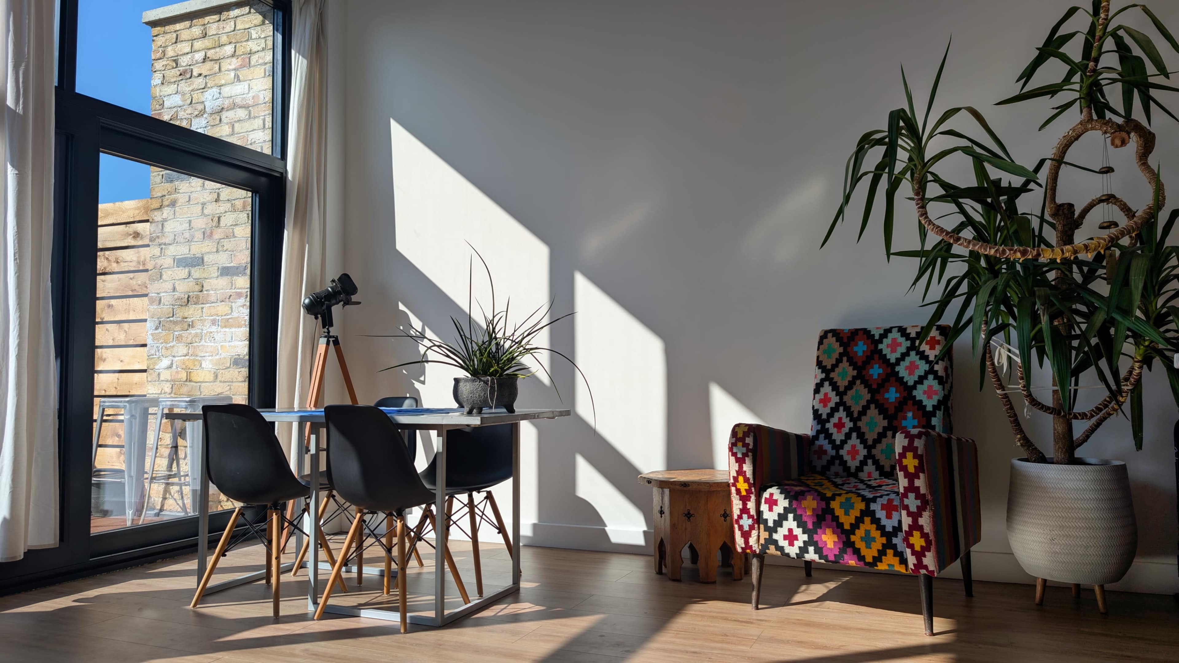 A brightly lit room features a dining table with chairs, a patterned armchair, a potted plant, and large windows casting shadows across the floor.