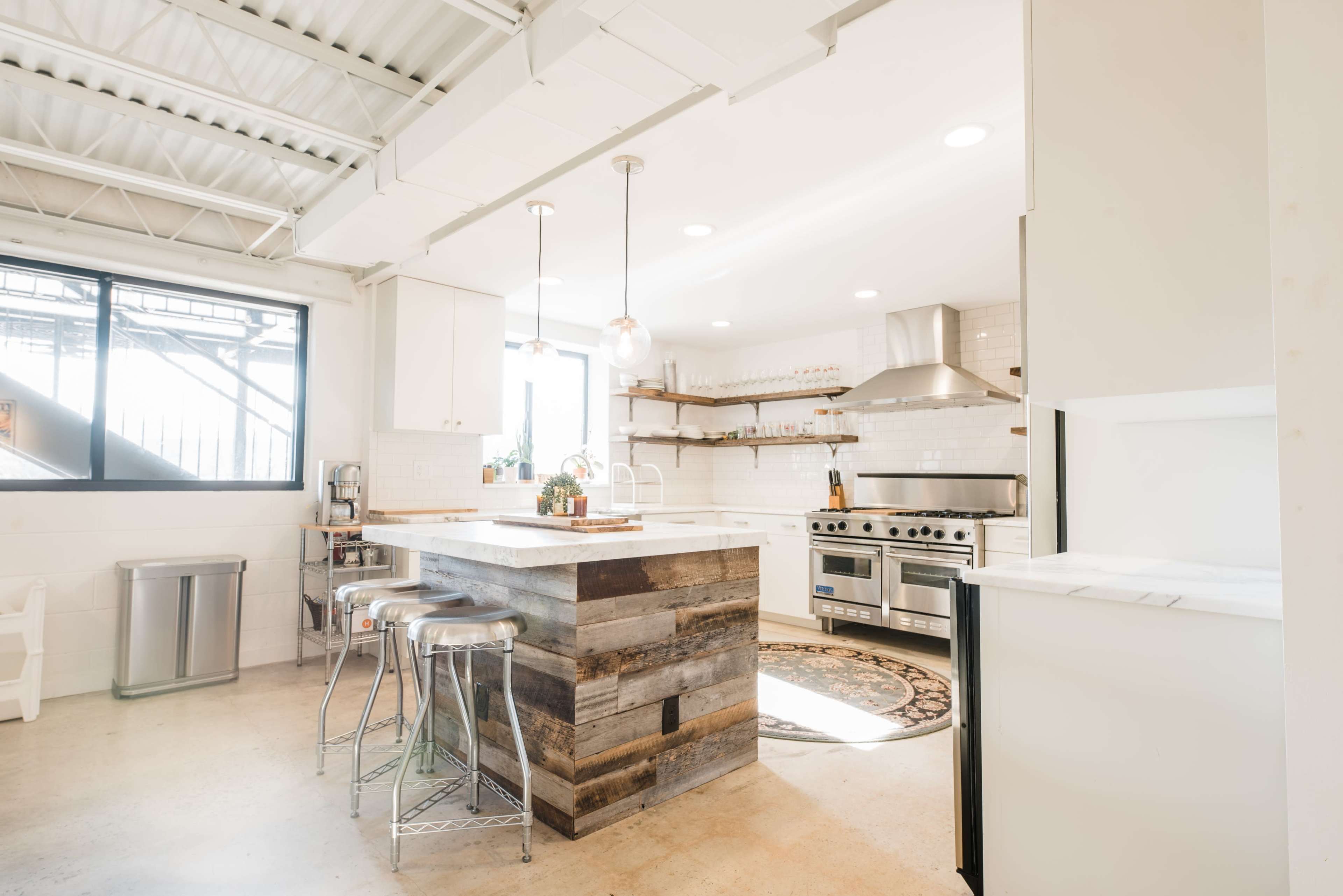 A modern kitchen features a spacious island made of reclaimed wood, stainless steel appliances, and large windows that allow natural light to enter.