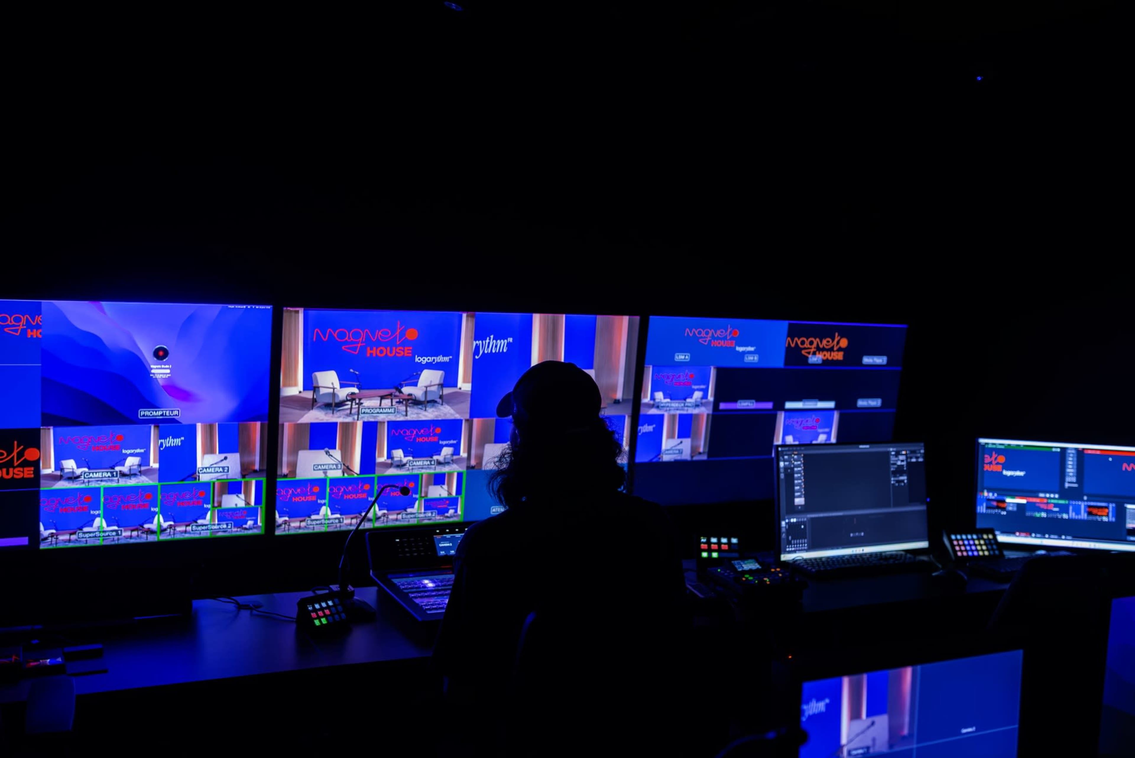 A person operates a video production setup featuring multiple monitors and control equipment in a dimly lit studio.
