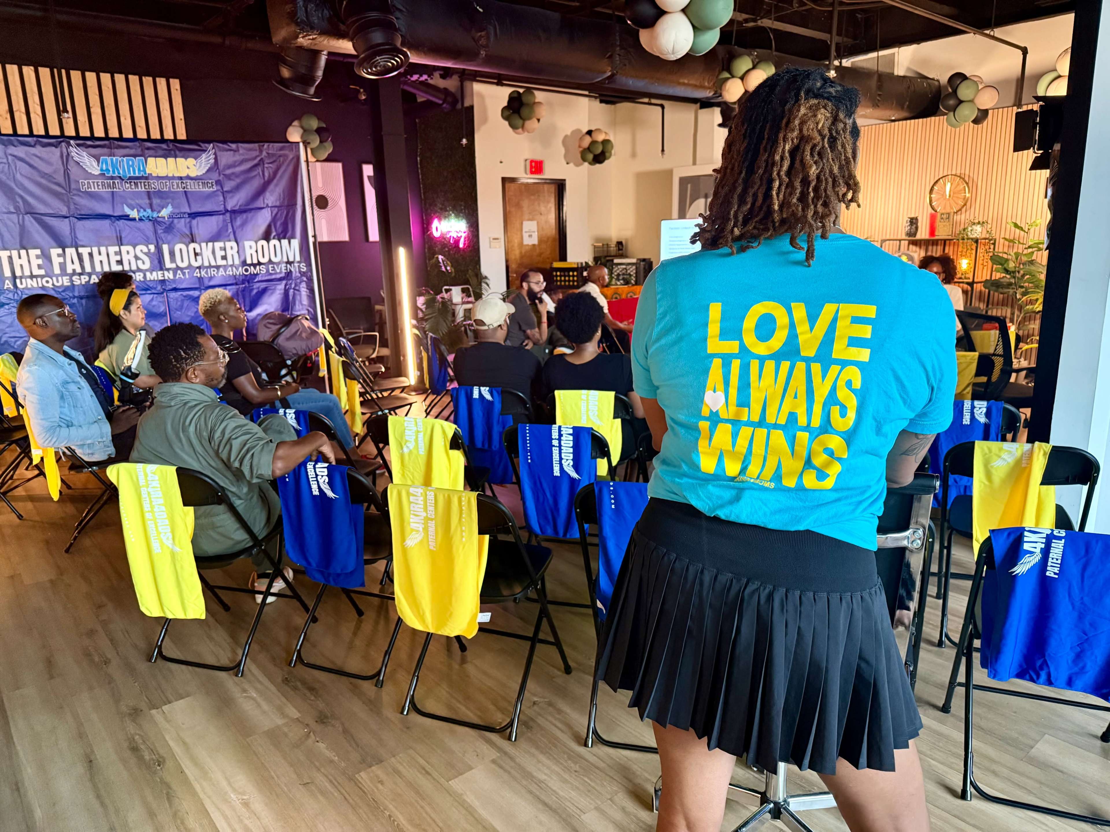 A speaker stands in front of an audience seated on colorful chairs, with a focus on the message "LOVE ALWAYS WINS" printed on the back of their shirt.