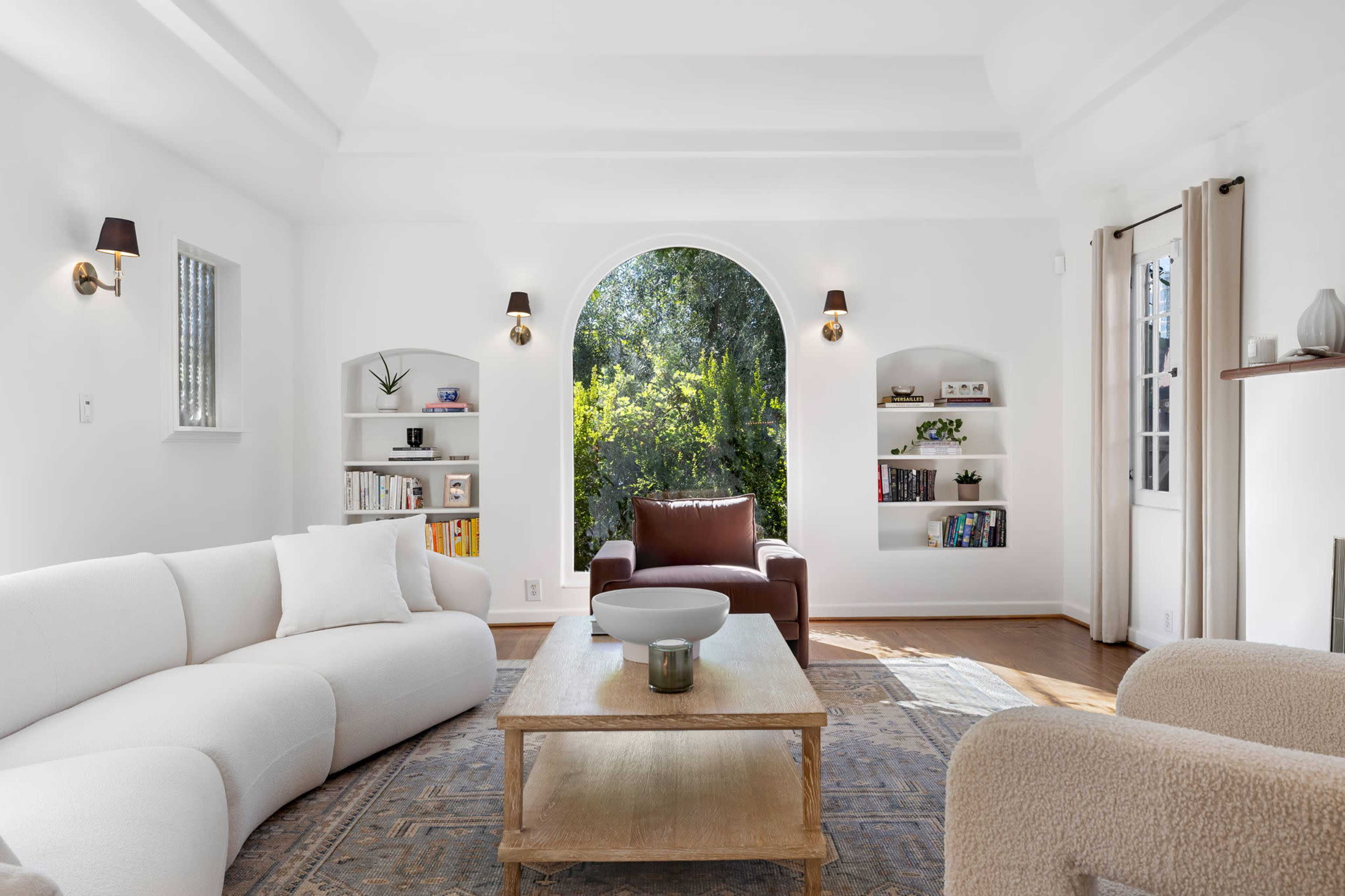 A bright living room featuring a curved white sofa, a brown leather armchair, a wooden coffee table, and shelves filled with books, all set against a large arched window that opens to a view of greenery.