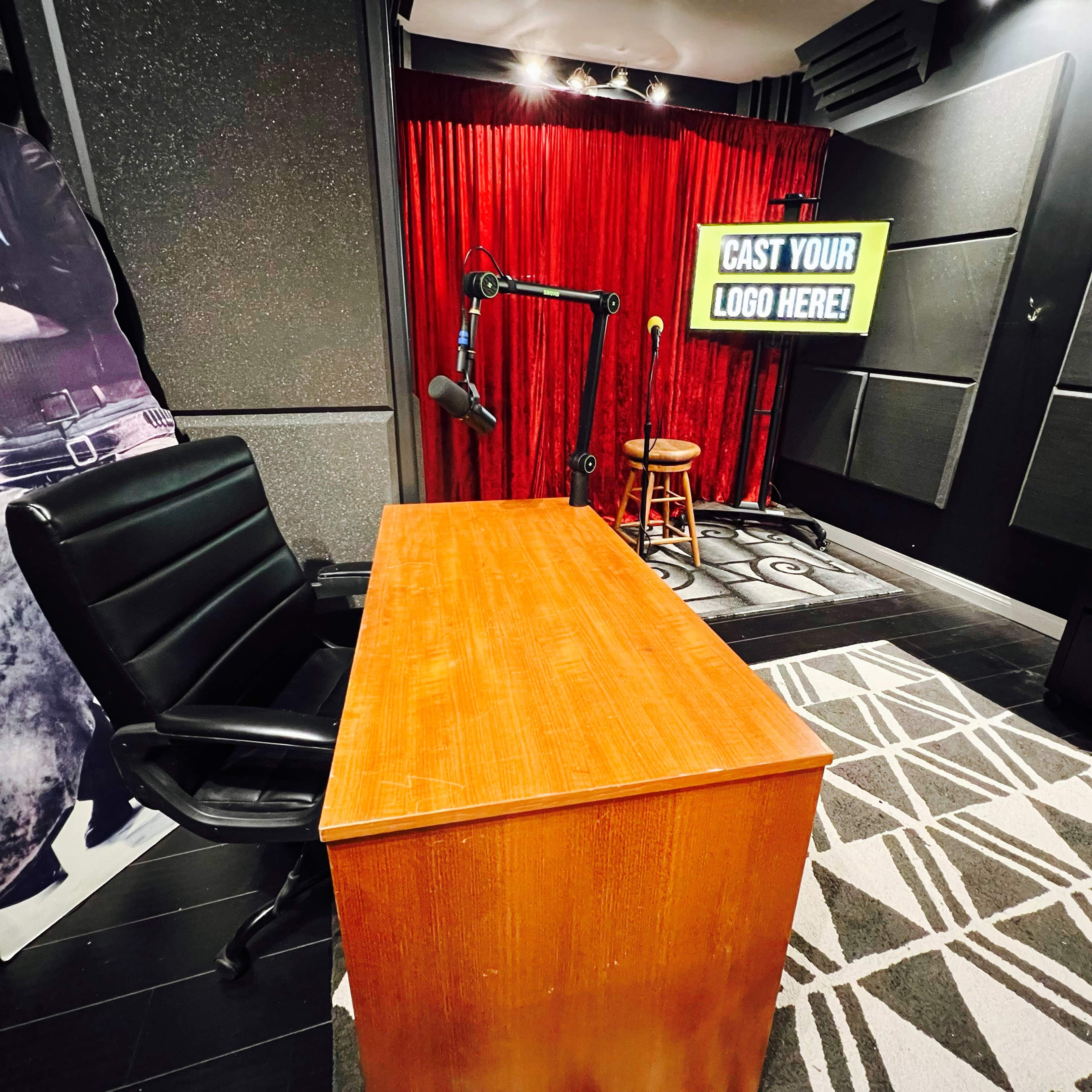 The image shows a dimly lit podcasting studio featuring a wooden desk and an adjustable microphone, with a stool and a large screen displaying the text "CAST YOUR LOGO HERE!" in the background.