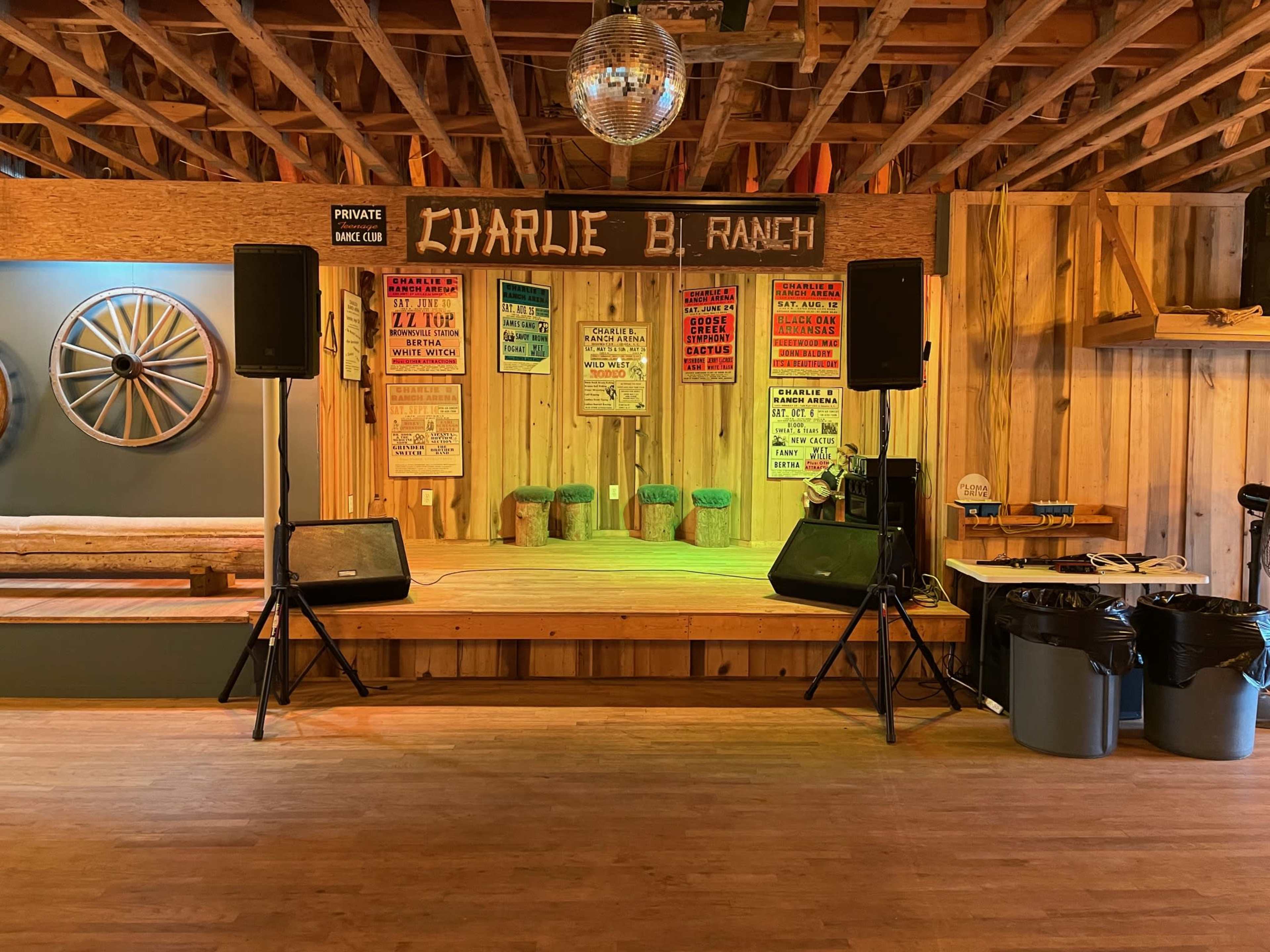 A wooden stage at the Charlie B. Ranch, equipped with speakers and a disco ball, decorated with vintage concert posters.