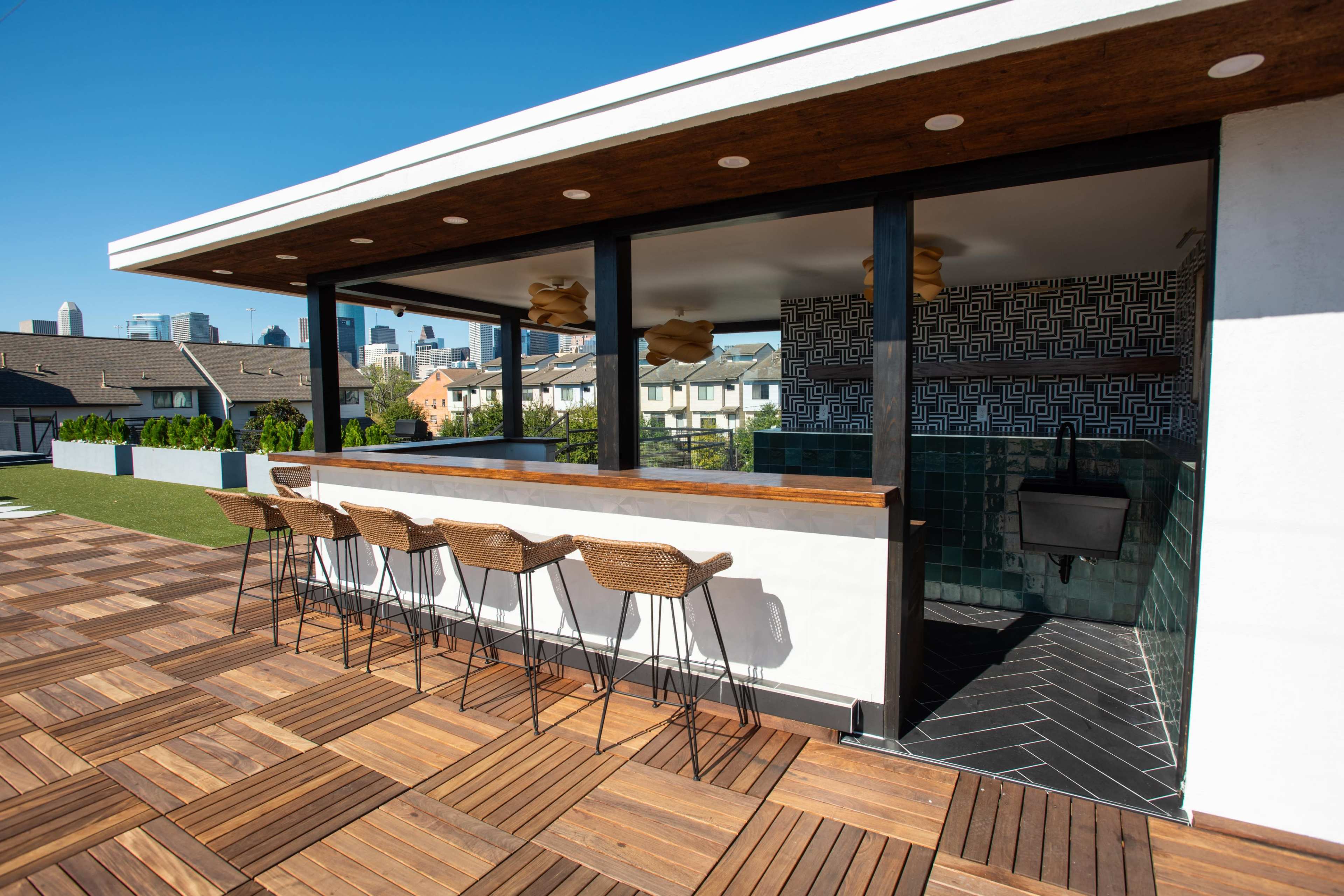 A modern rooftop bar featuring bar stools lined up against a wooden counter with a decorative backsplash and an open view of the city skyline.