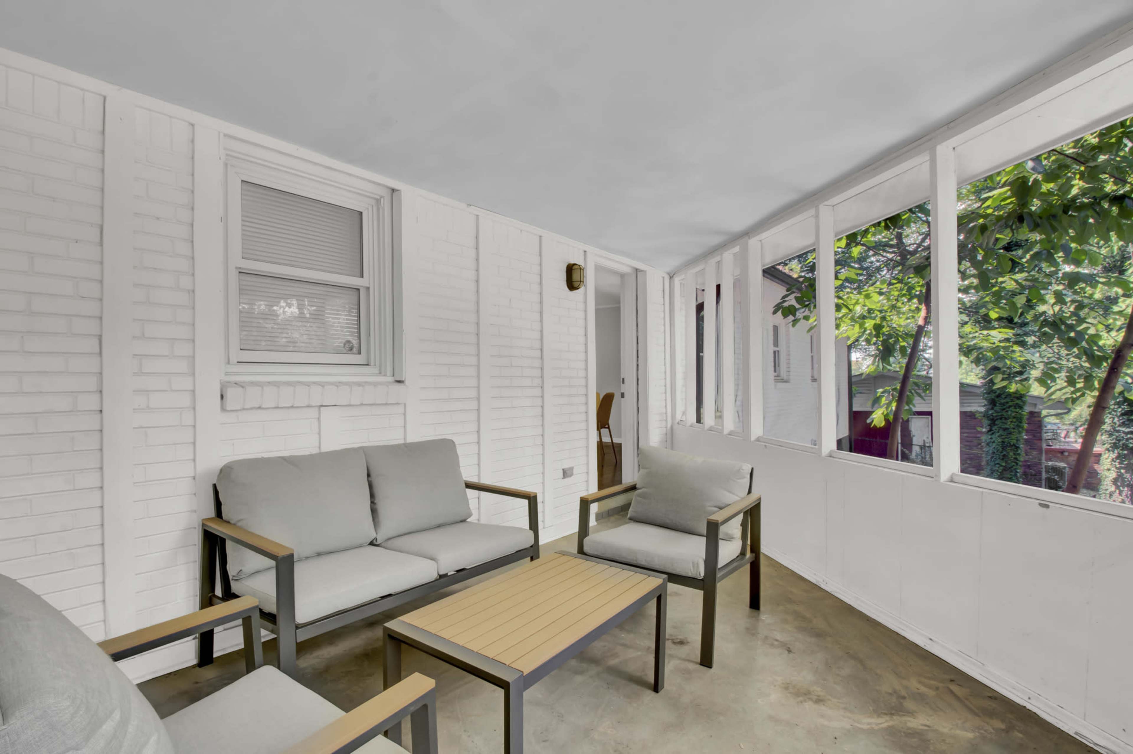 The image shows a screened-in porch featuring two gray armchairs, a coffee table, and a view of greenery outside.