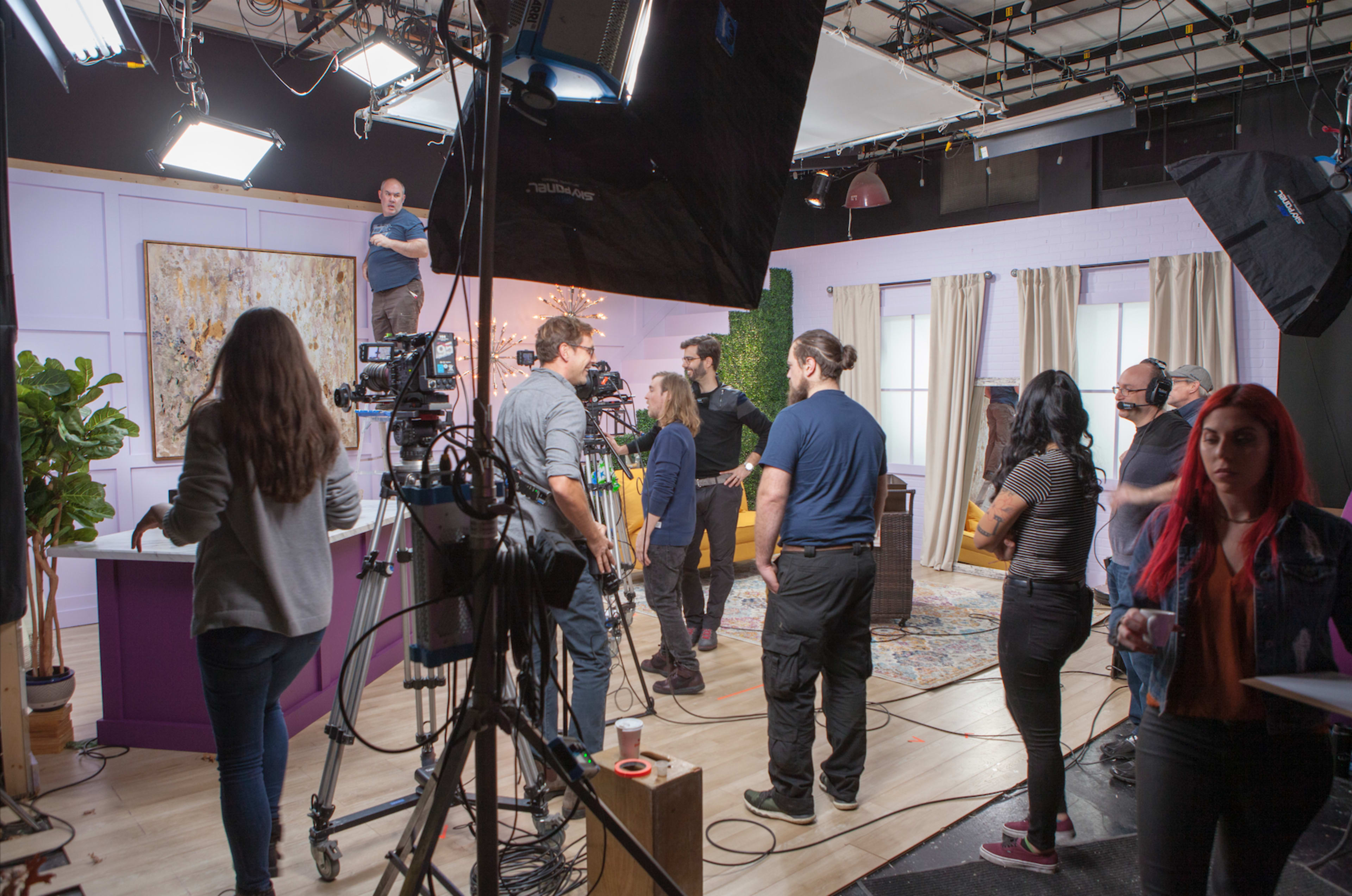 A film crew prepares for a shoot in a television studio with cameras, lighting equipment, and a backdrop arrangement.