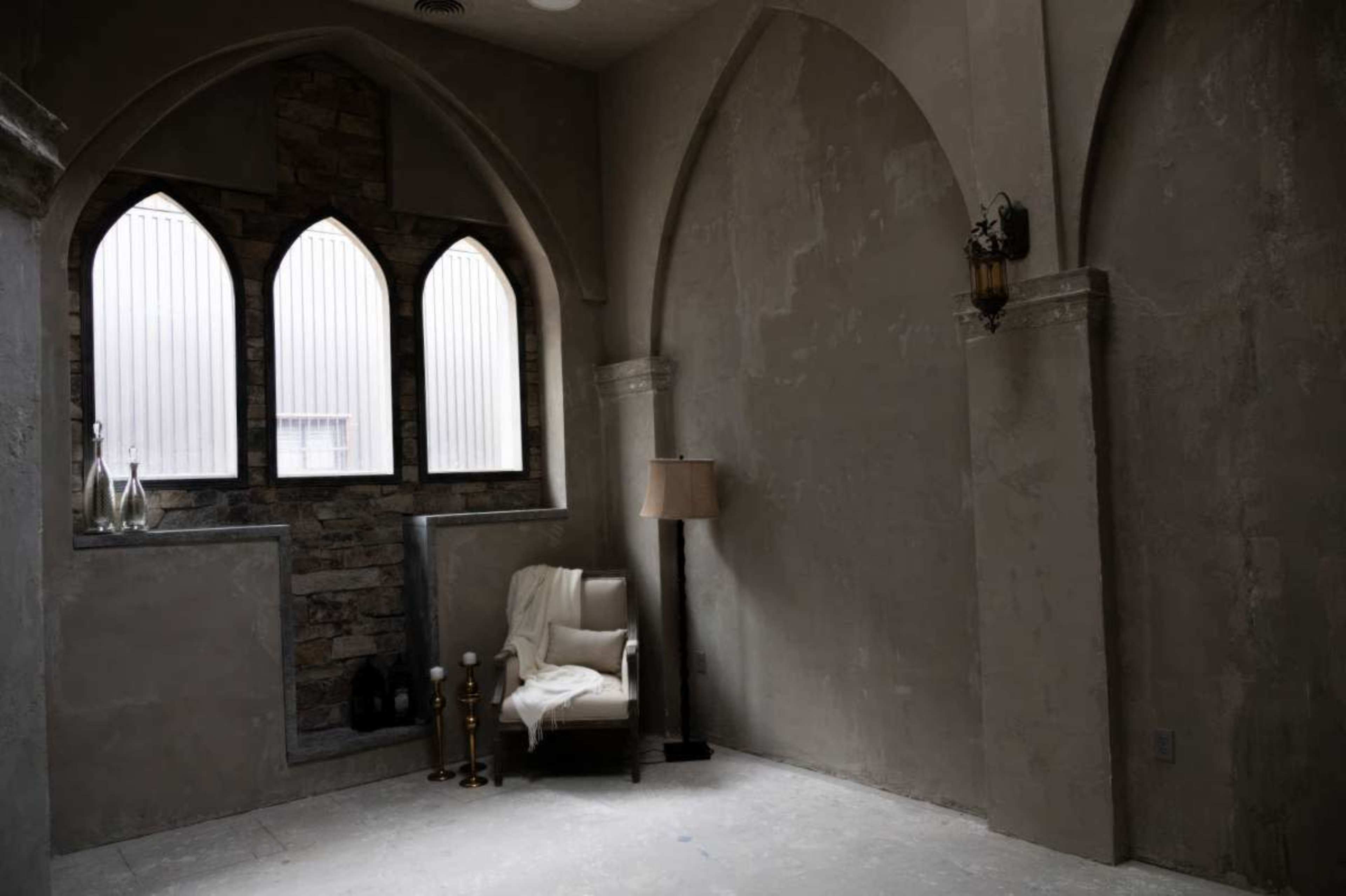 The image shows an interior corner of a room featuring a chair beside a lamp, with arched windows and stone accents on the walls.