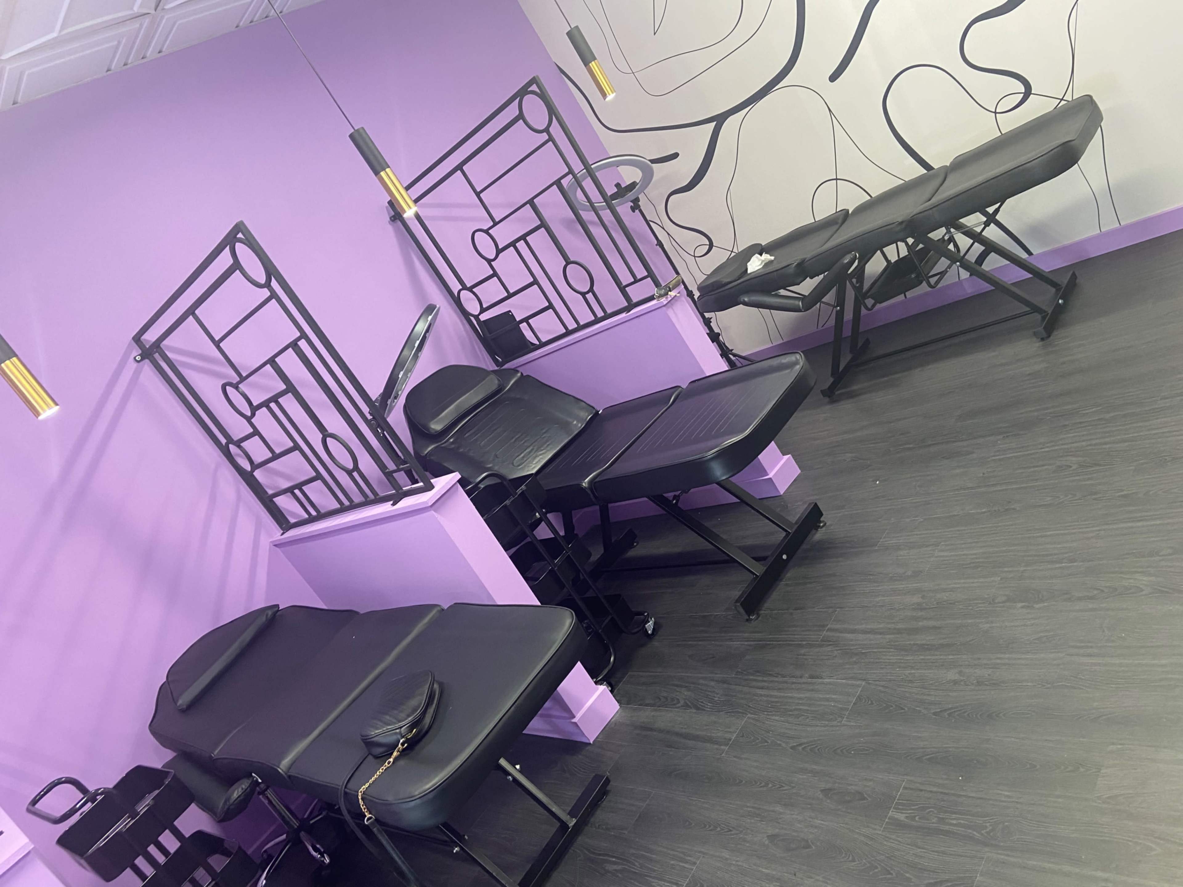 The image shows a clean beauty treatment room with two black treatment tables against a purple wall featuring minimalist line art.