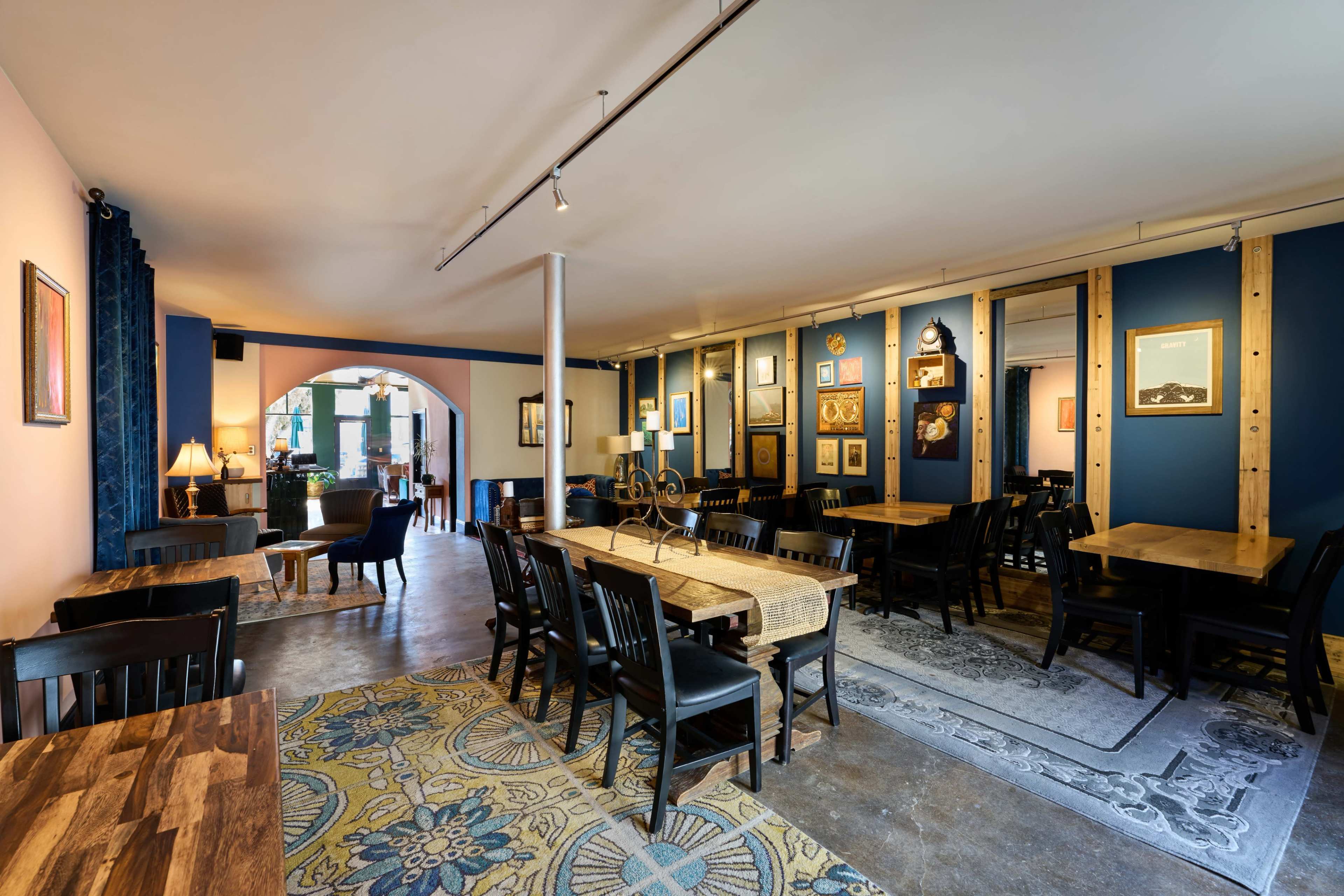 The image shows a modern café interior with a mix of black wooden tables and chairs, decorative artwork on the walls, and an area rug on the floor.