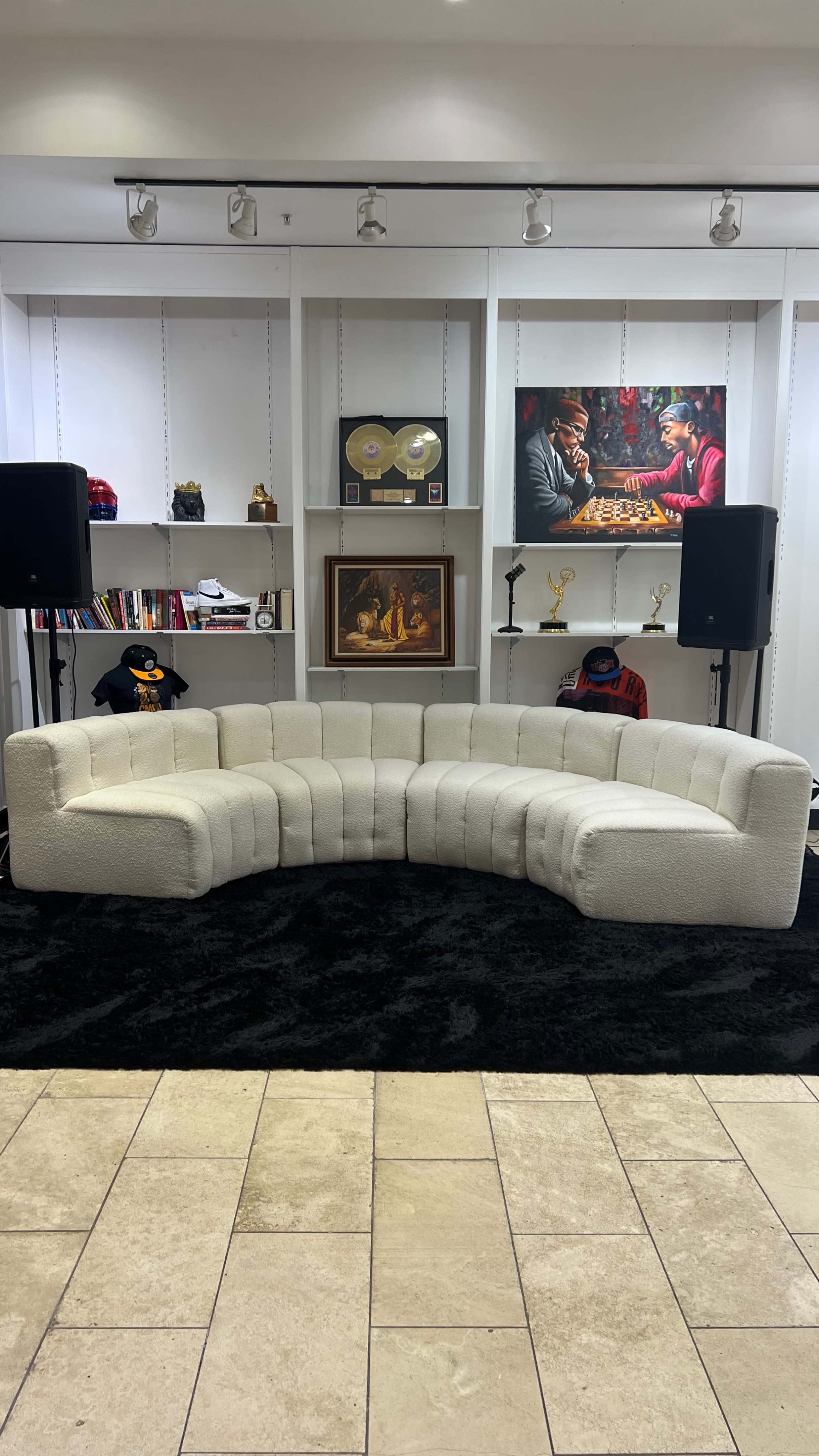 The image shows a curved white sofa placed on a black rug in a room featuring shelves with various decorations and two speakers on either side.