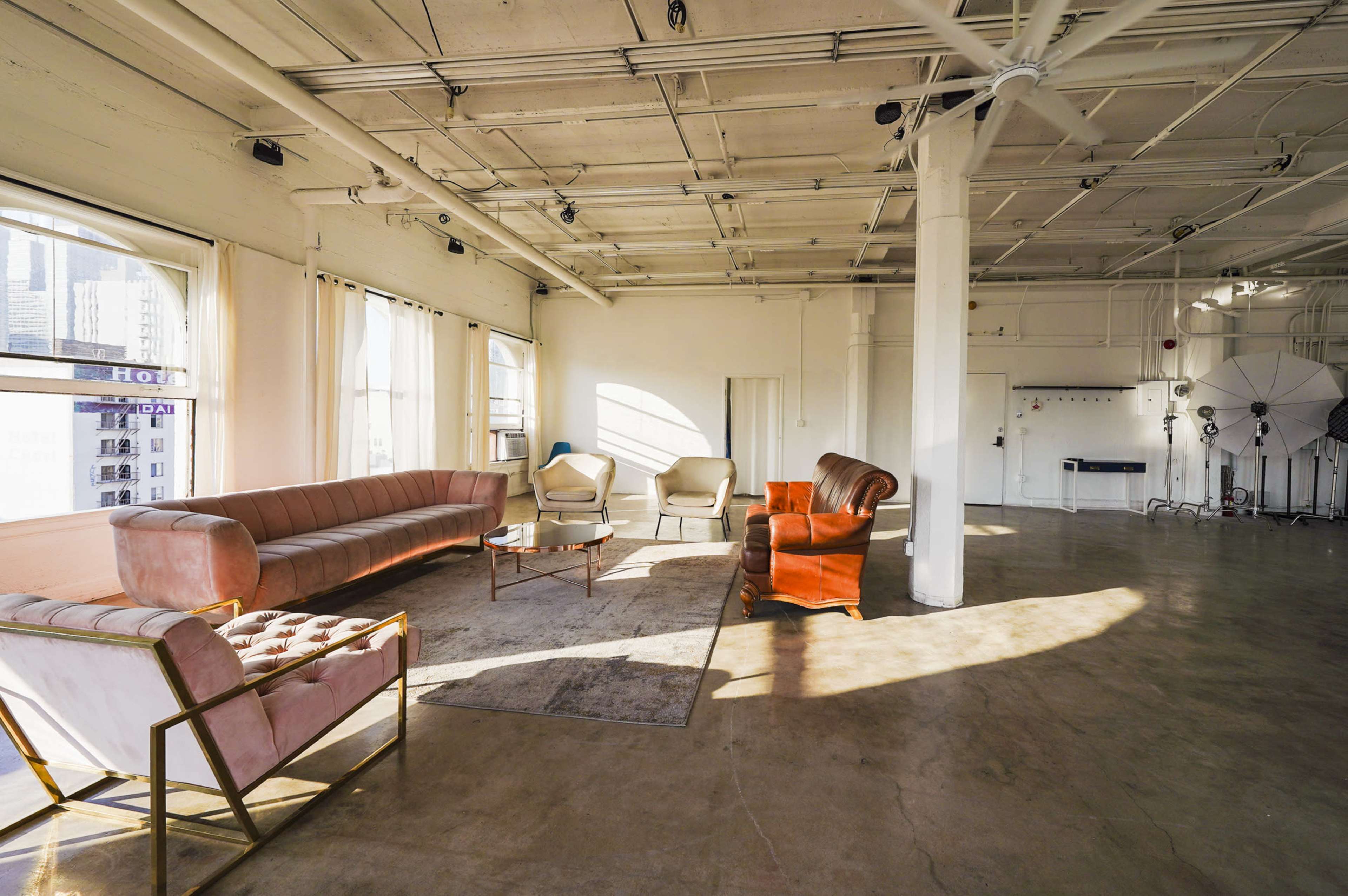 A spacious, well-lit lounge area with a mix of vintage and modern furniture, including a long pink sofa, a white armchair, and an orange armchair, set against a backdrop of large windows.