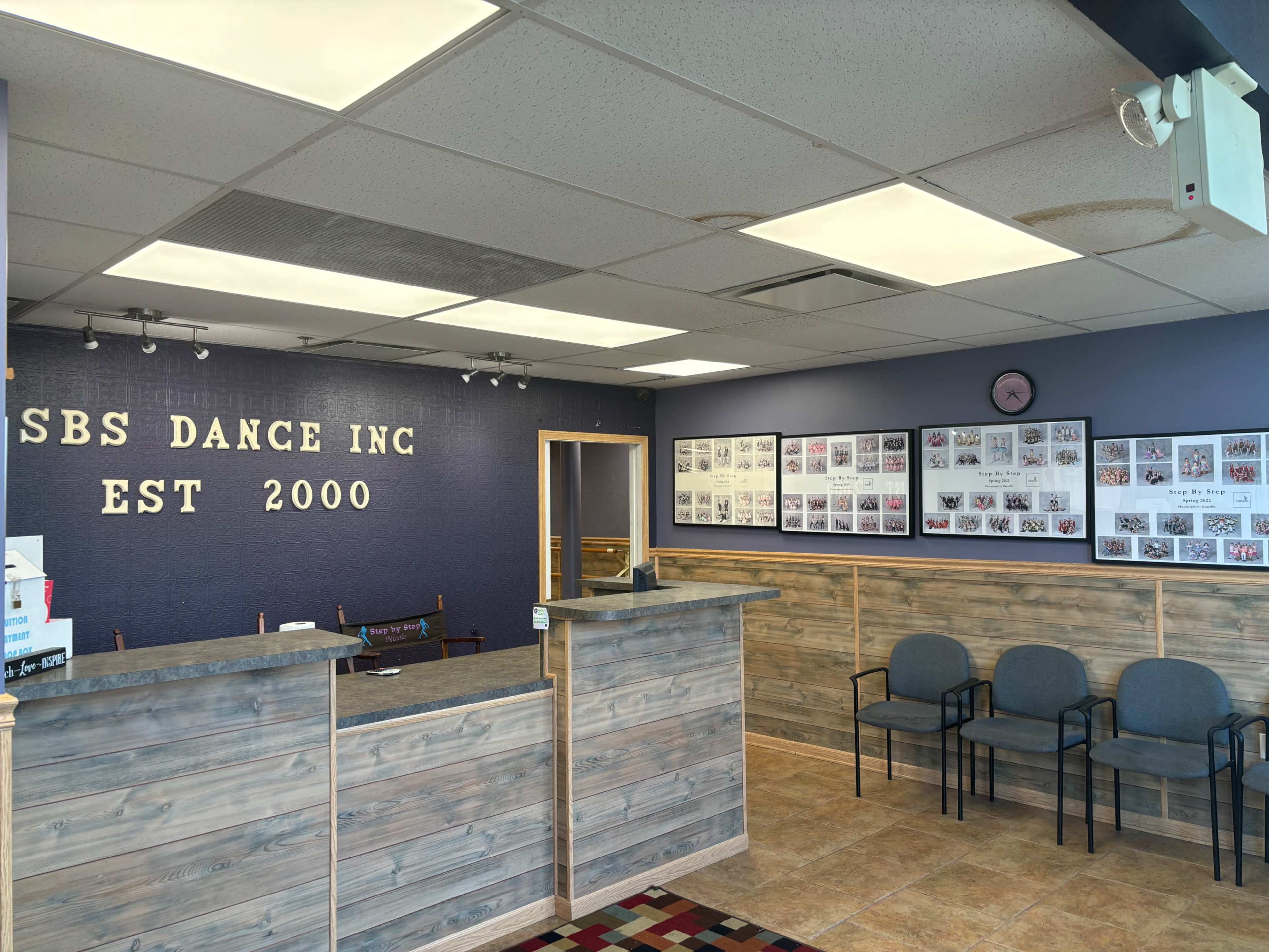 A reception area for SBS Dance Inc., featuring a wooden front desk, gray chairs, and framed photographs on the walls.