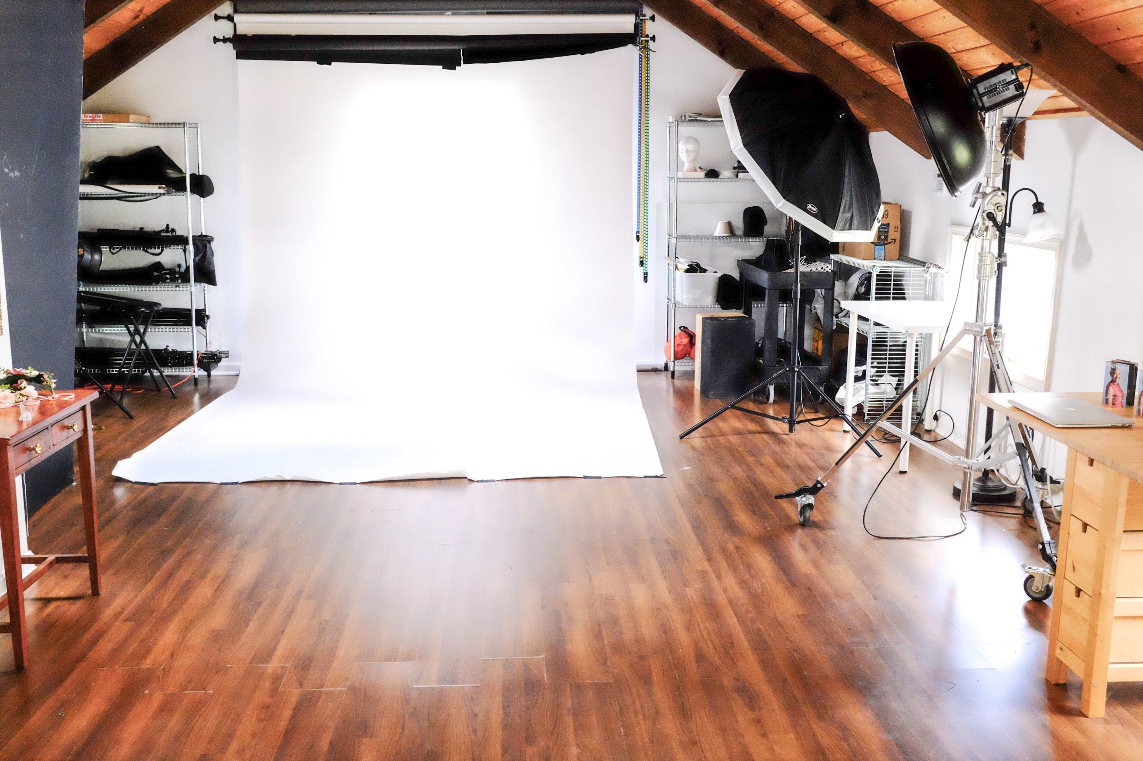 A bright photography studio with a white backdrop, wooden flooring, and various lighting equipment arranged around the space.