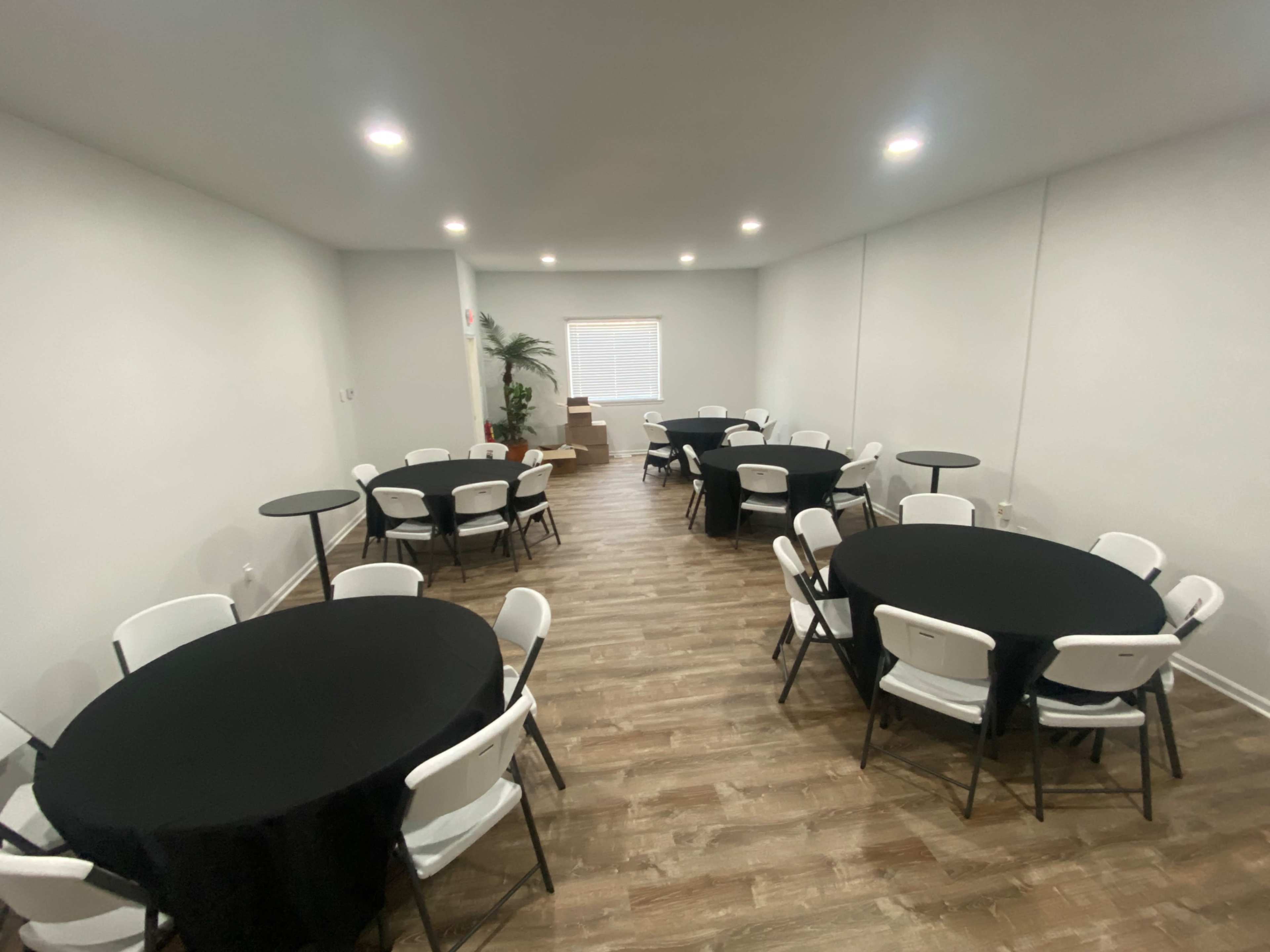 The image shows a spacious room set up with several round tables covered in black tablecloths, surrounded by white chairs, and lit by ceiling lights.