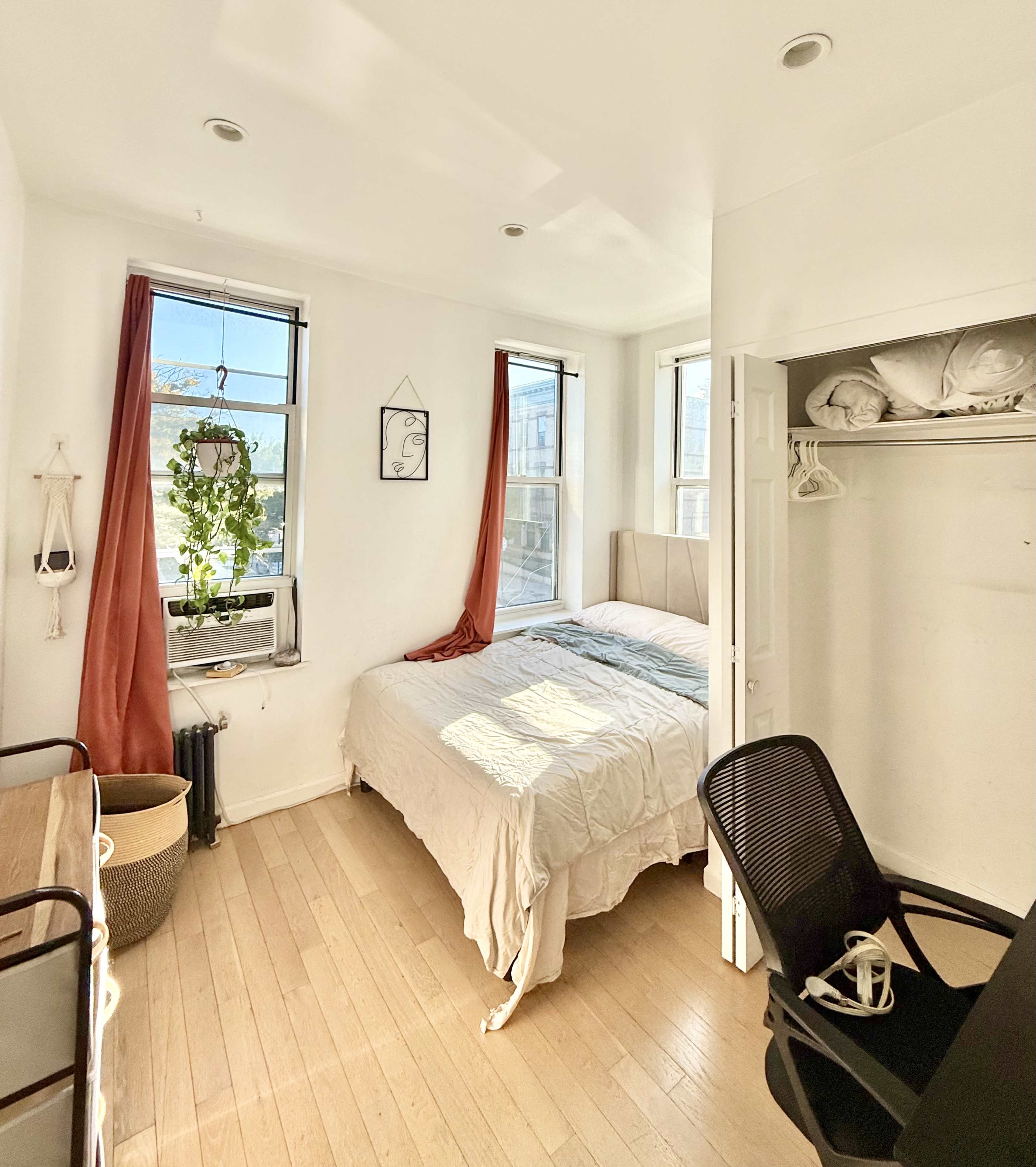 The image shows a bright bedroom featuring a bed, windows with sheer curtains, a desk with a chair, and a closet with visible linens.