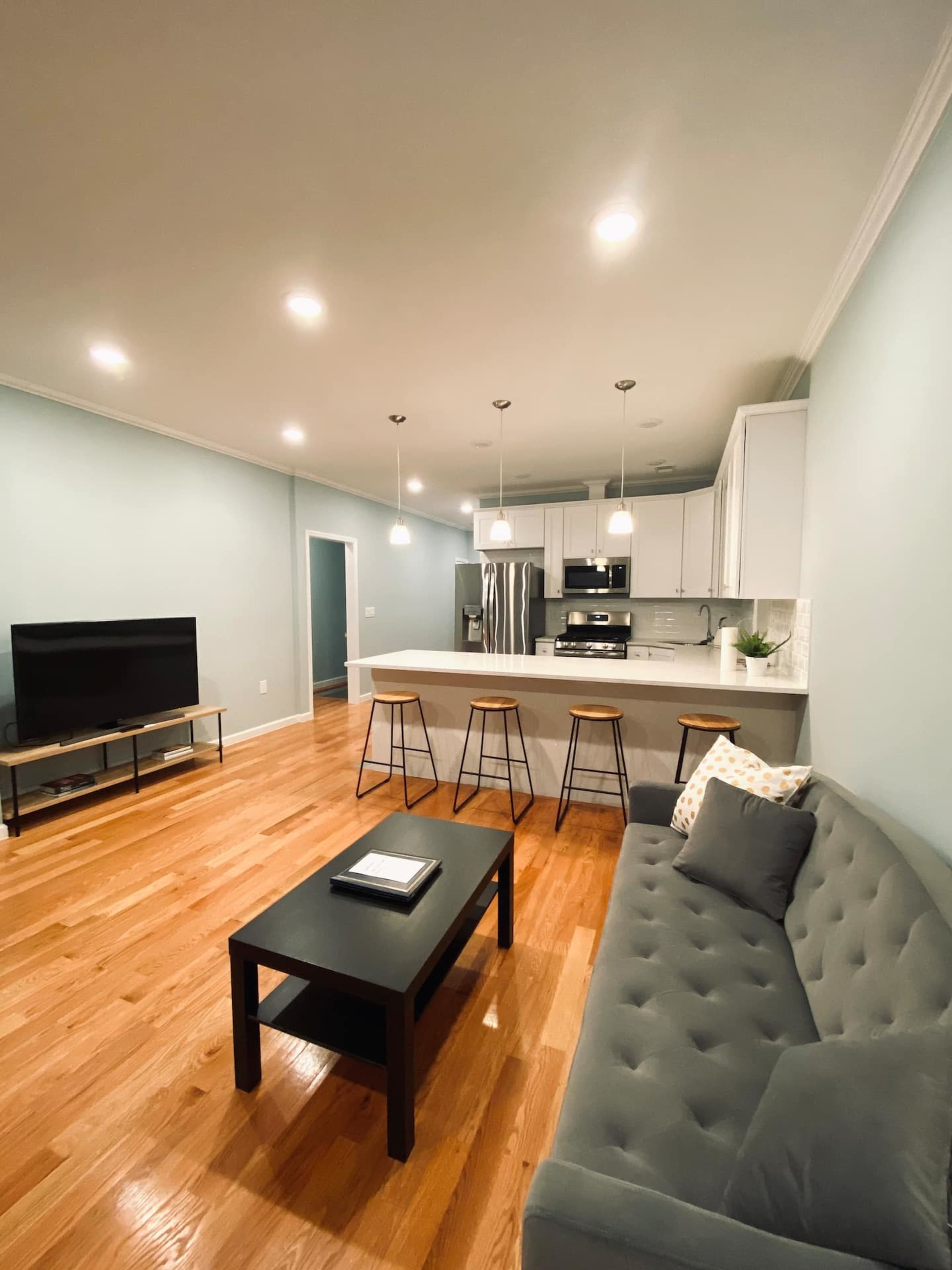 A modern living room and kitchen area featuring a gray couch, a black coffee table, bar stools at a kitchen island, and a large television on a stand.