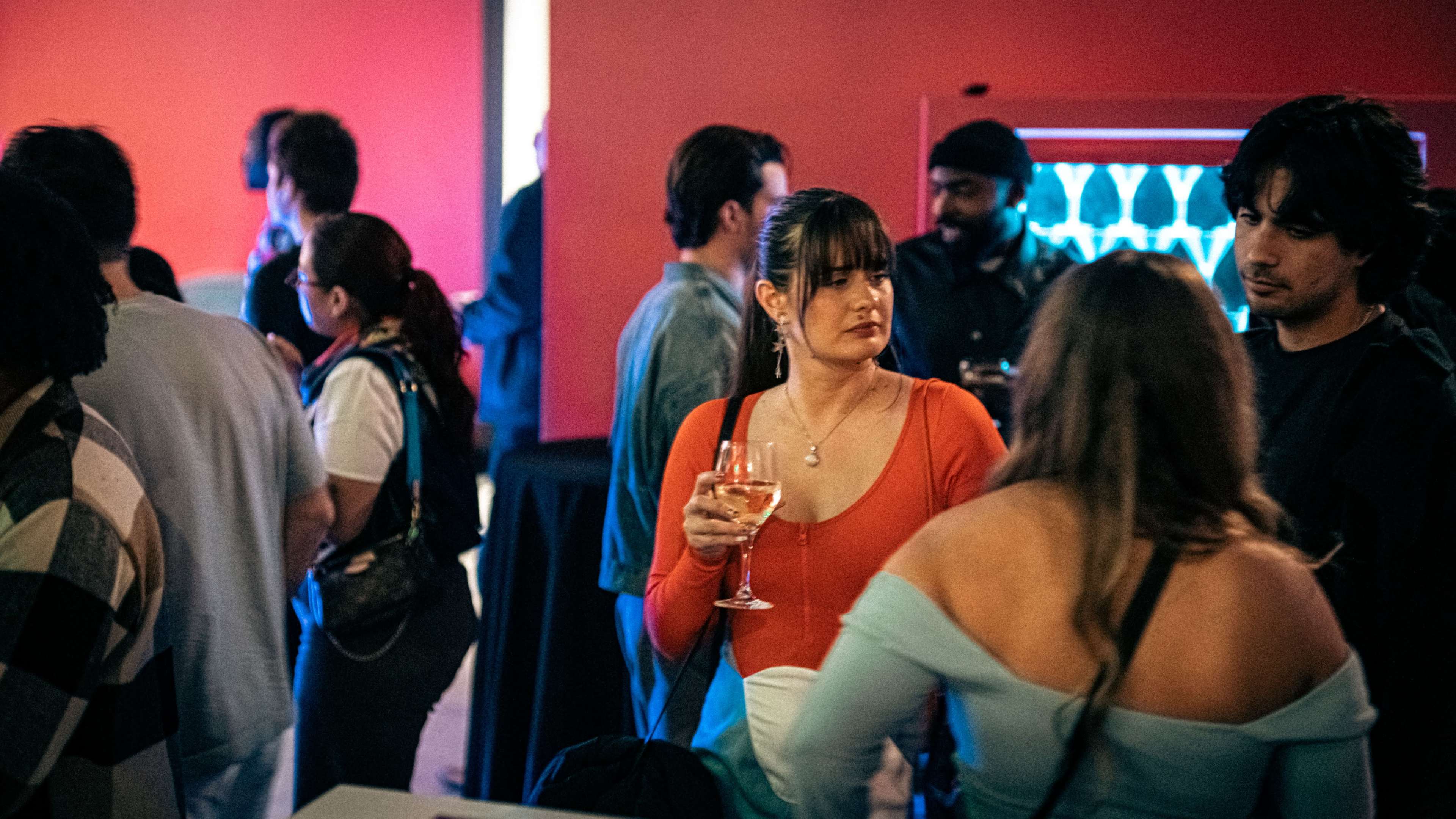 A crowded social event features a group of people mingling, with one woman holding a drink and wearing an orange top in the foreground.