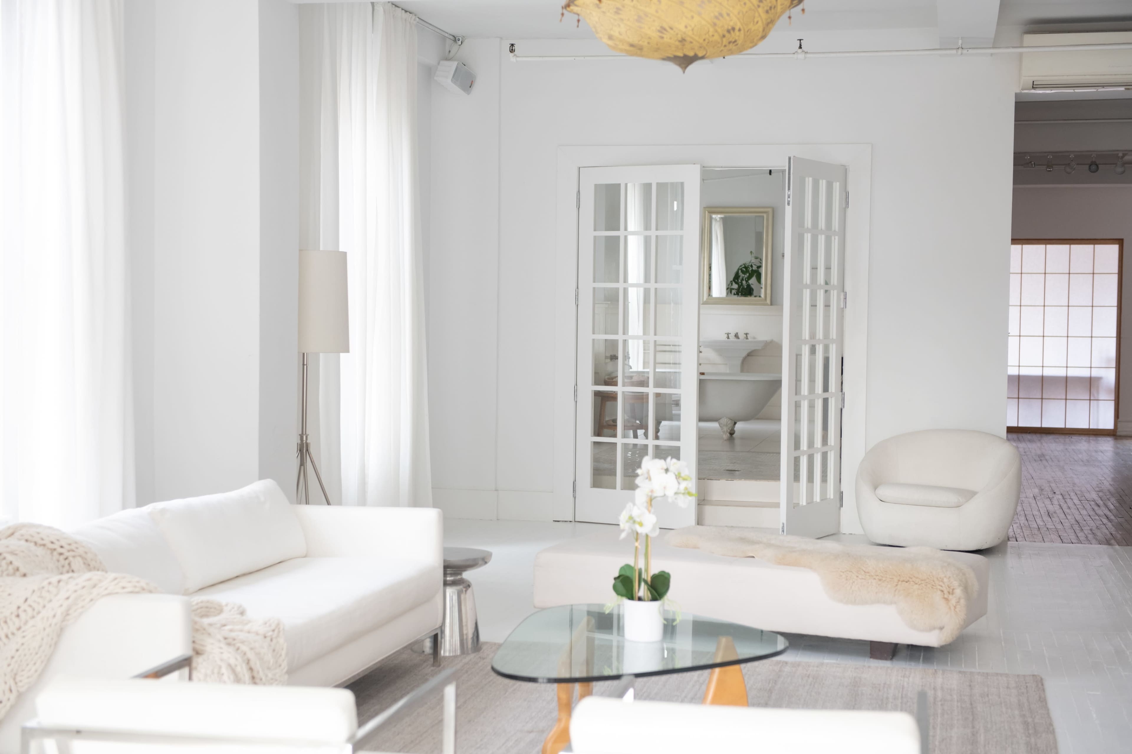 A bright, modern living space featuring white furniture, a glass coffee table, and an open door leading to a bathroom area.