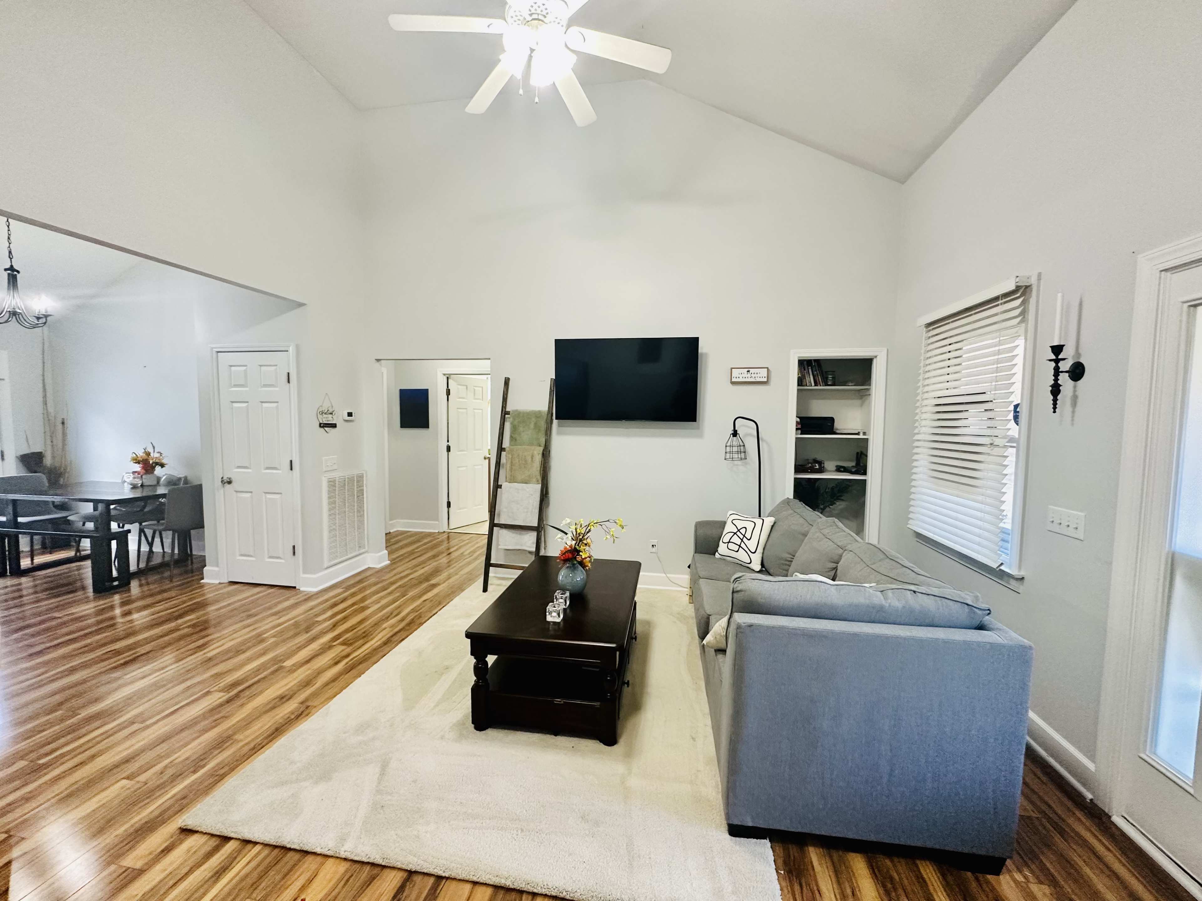 A spacious living room with a gray sofa, a dark coffee table, and a flat-screen television mounted on a light-colored wall.