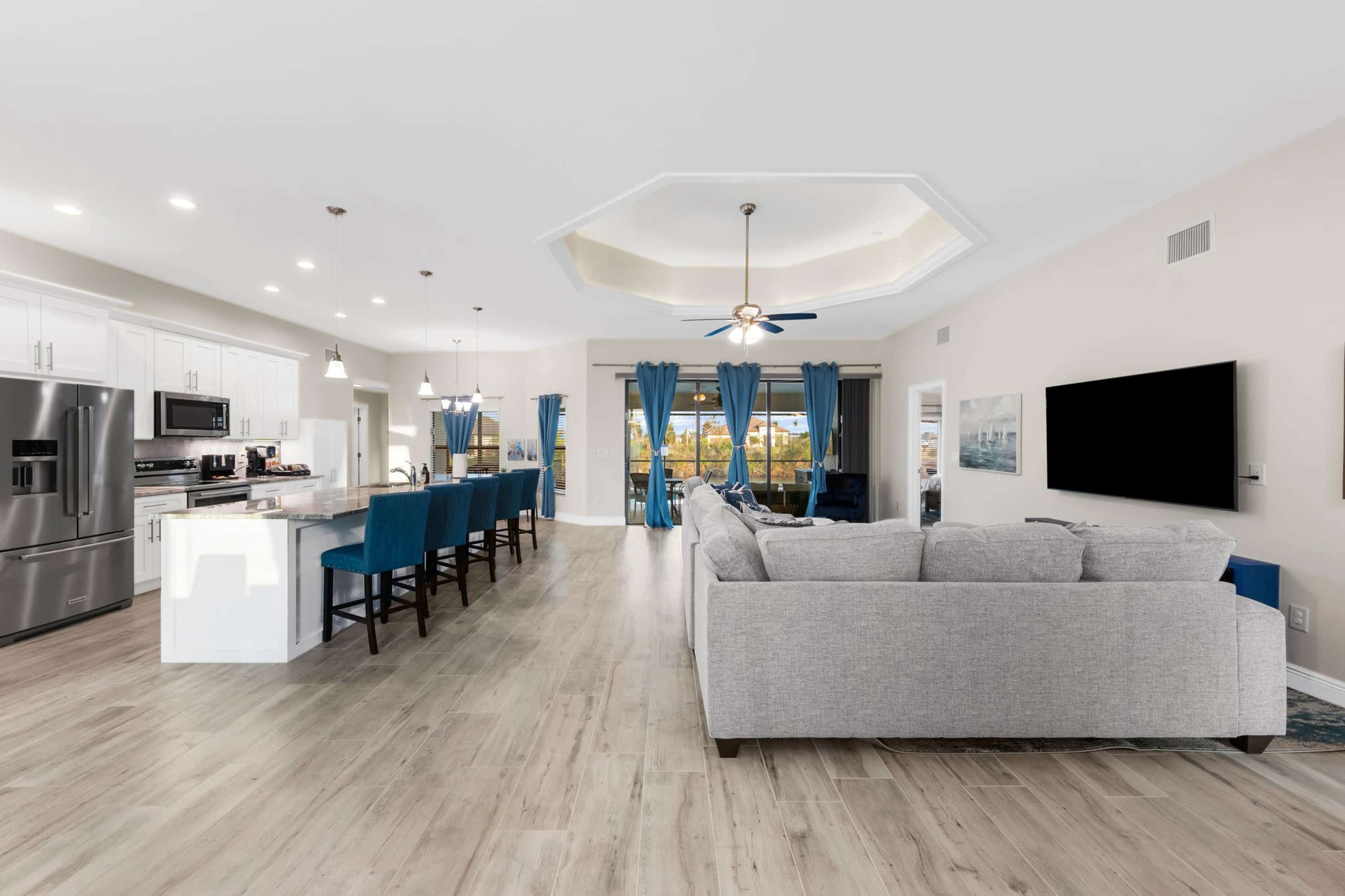A bright, open living space with a gray sectional sofa, a kitchen with white cabinets, and blue bar stools.