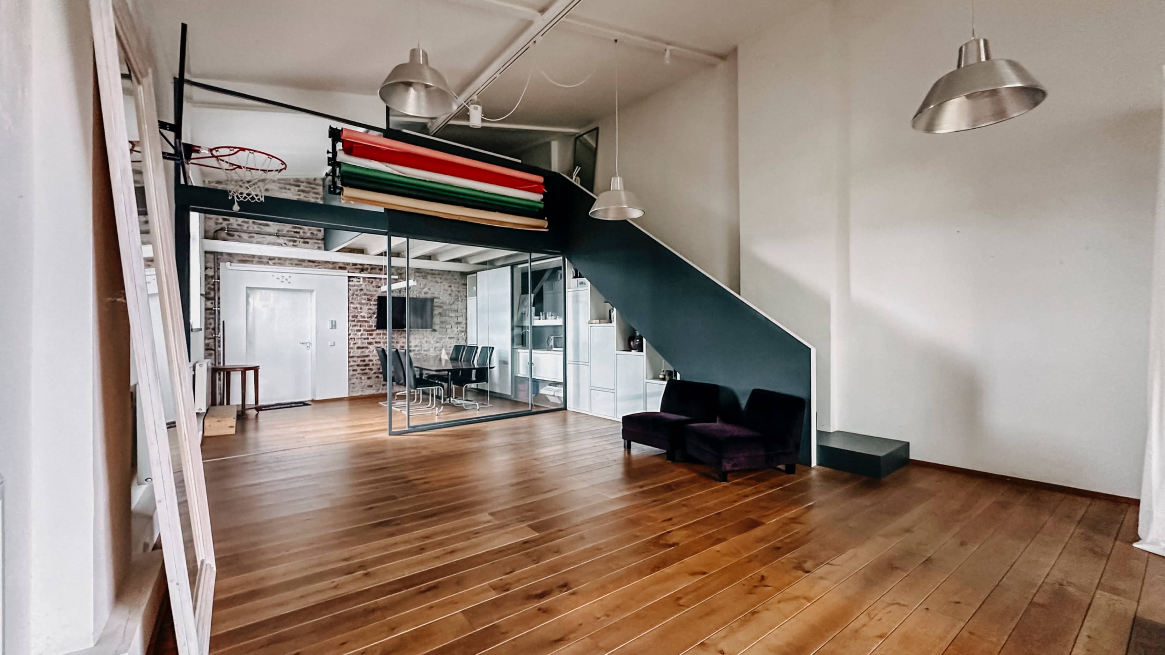 The image shows an open, modern space featuring wooden floors, an industrial-style staircase, a basketball hoop, and a glass-walled room with chairs and a light fixture.
