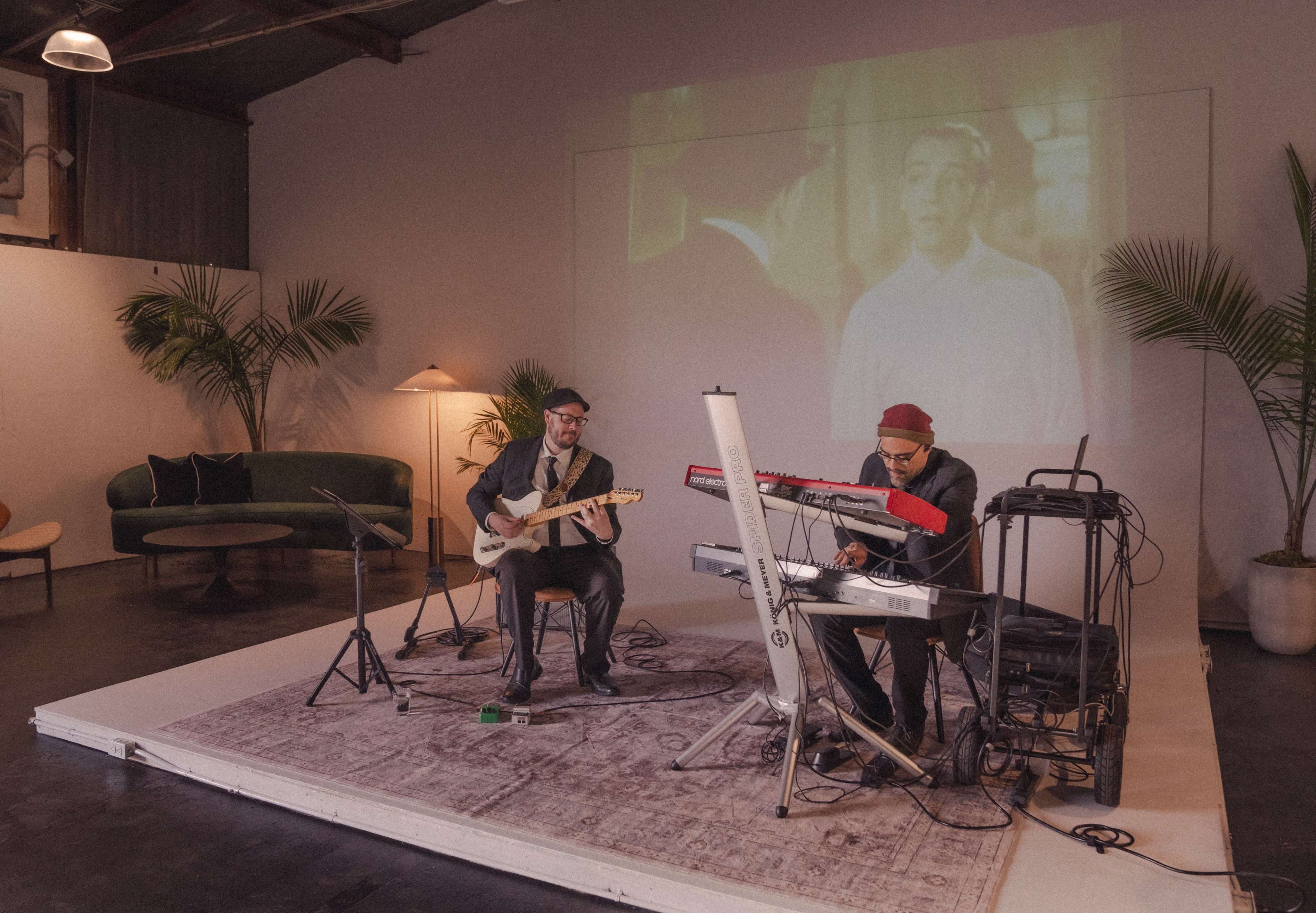 Two musicians perform on a small stage in a modern space with a projected film in the background.