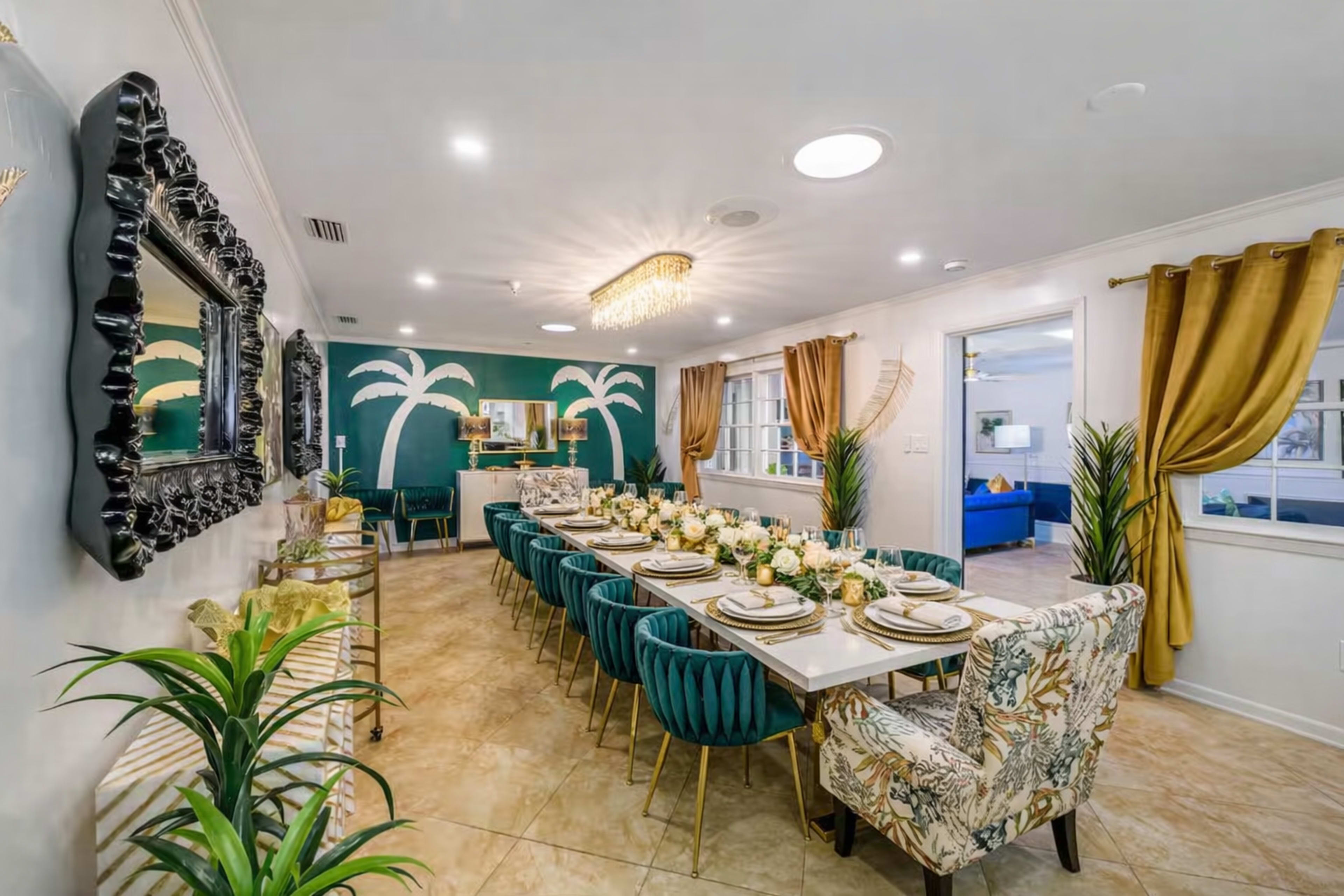 A long dining table is set with decorative tableware and surrounded by patterned chairs in a room featuring tropical-themed wall art and elegant lighting.