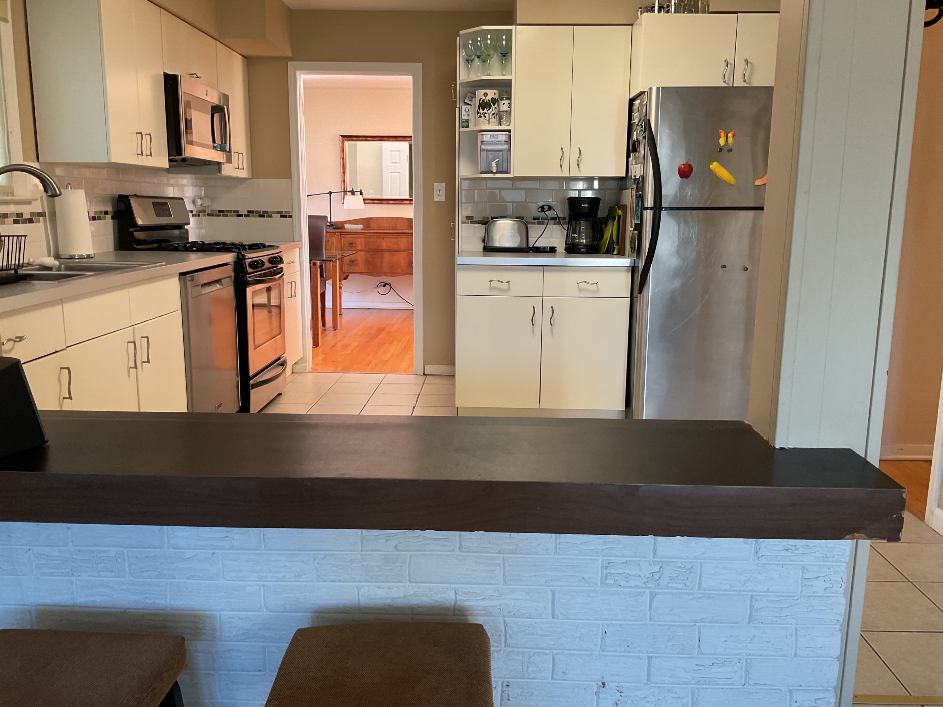 The image shows a kitchen with white cabinets, stainless steel appliances, and a bar counter that separates the kitchen from a small dining area visible in the background.