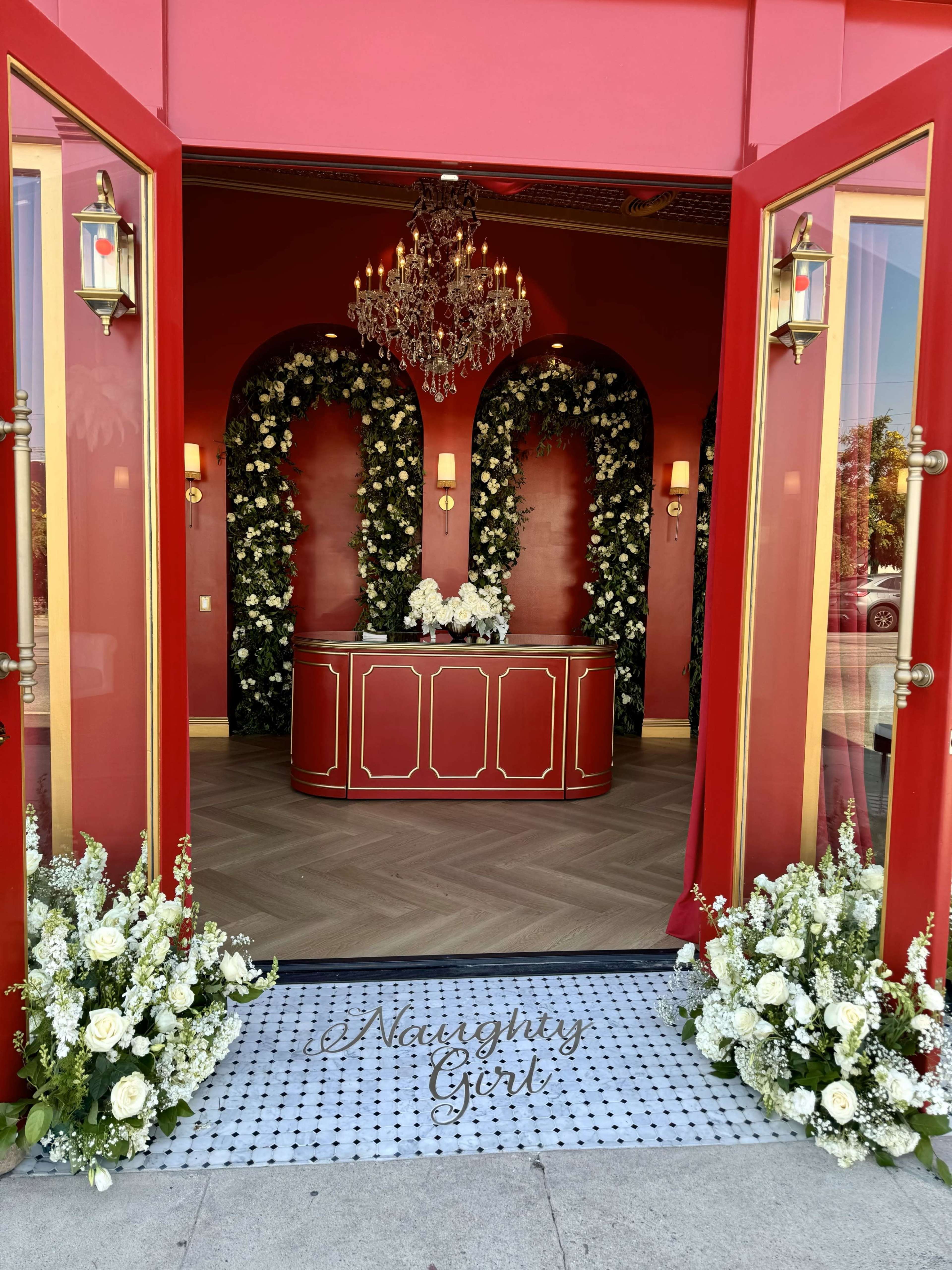 The entrance features a red facade with double doors flanked by floral arrangements and a glamorous chandelier inside.