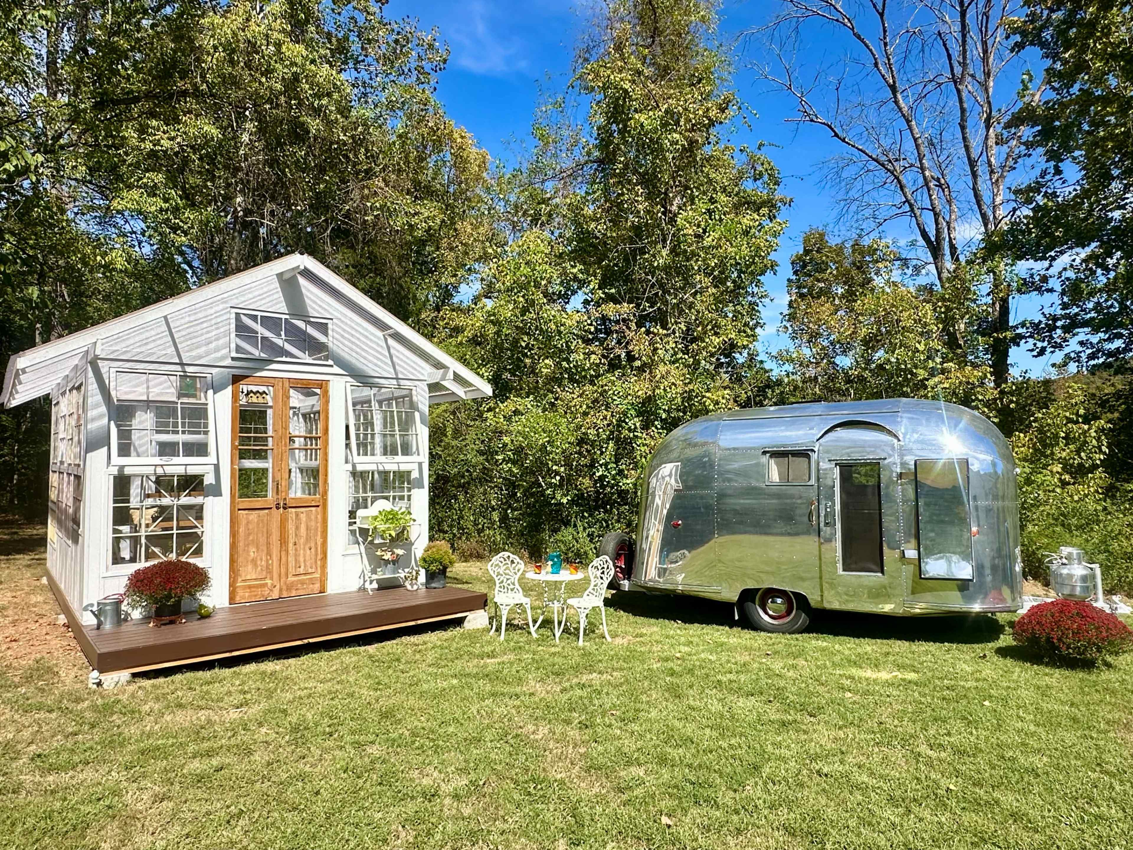 A shiny silver trailer is parked next to a small glass-fronted shed with a wooden deck, surrounded by greenery and blue sky.