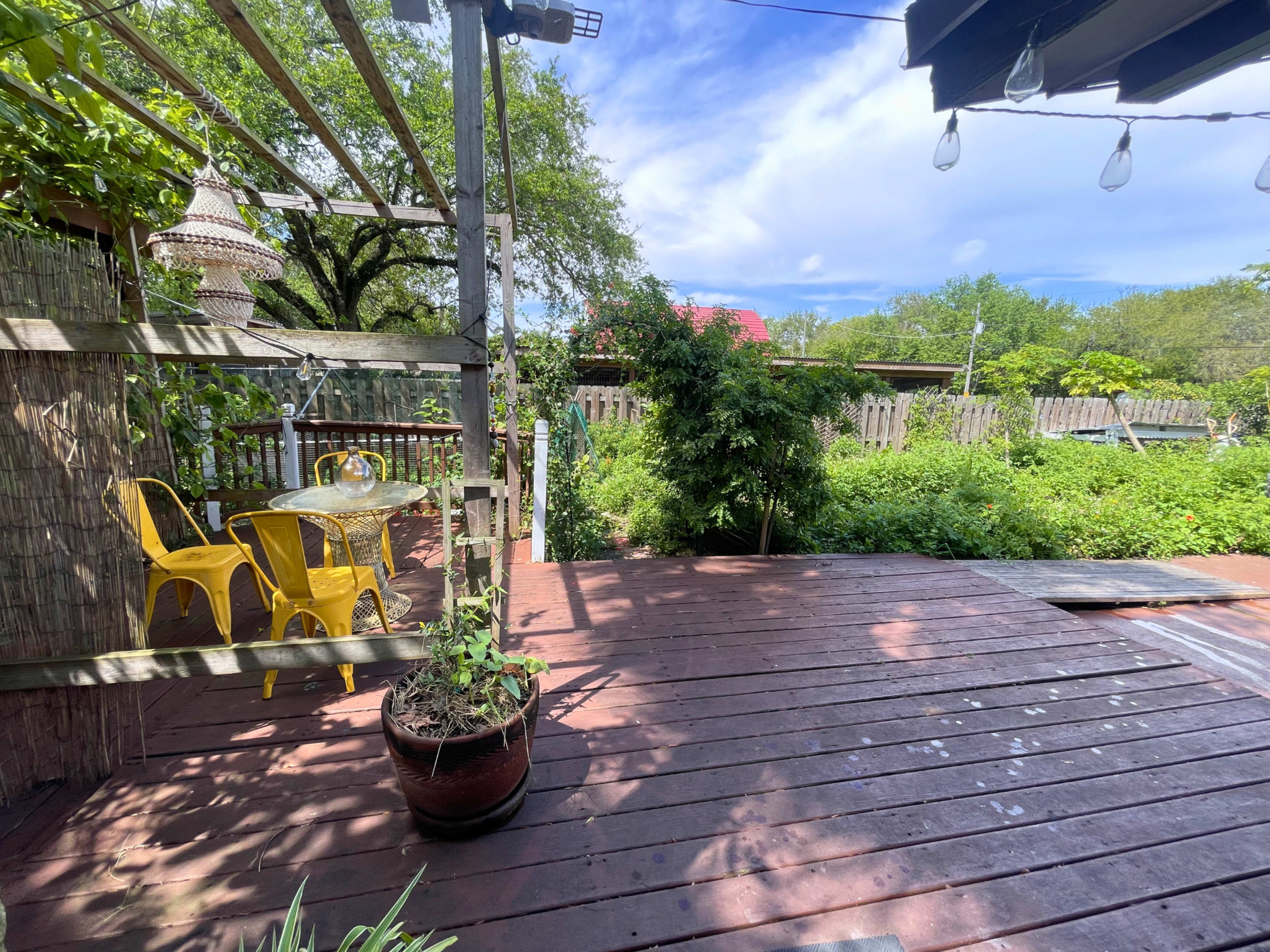The image shows a wooden deck with a small round glass table and yellow chairs, surrounded by greenery and under a partly cloudy sky.