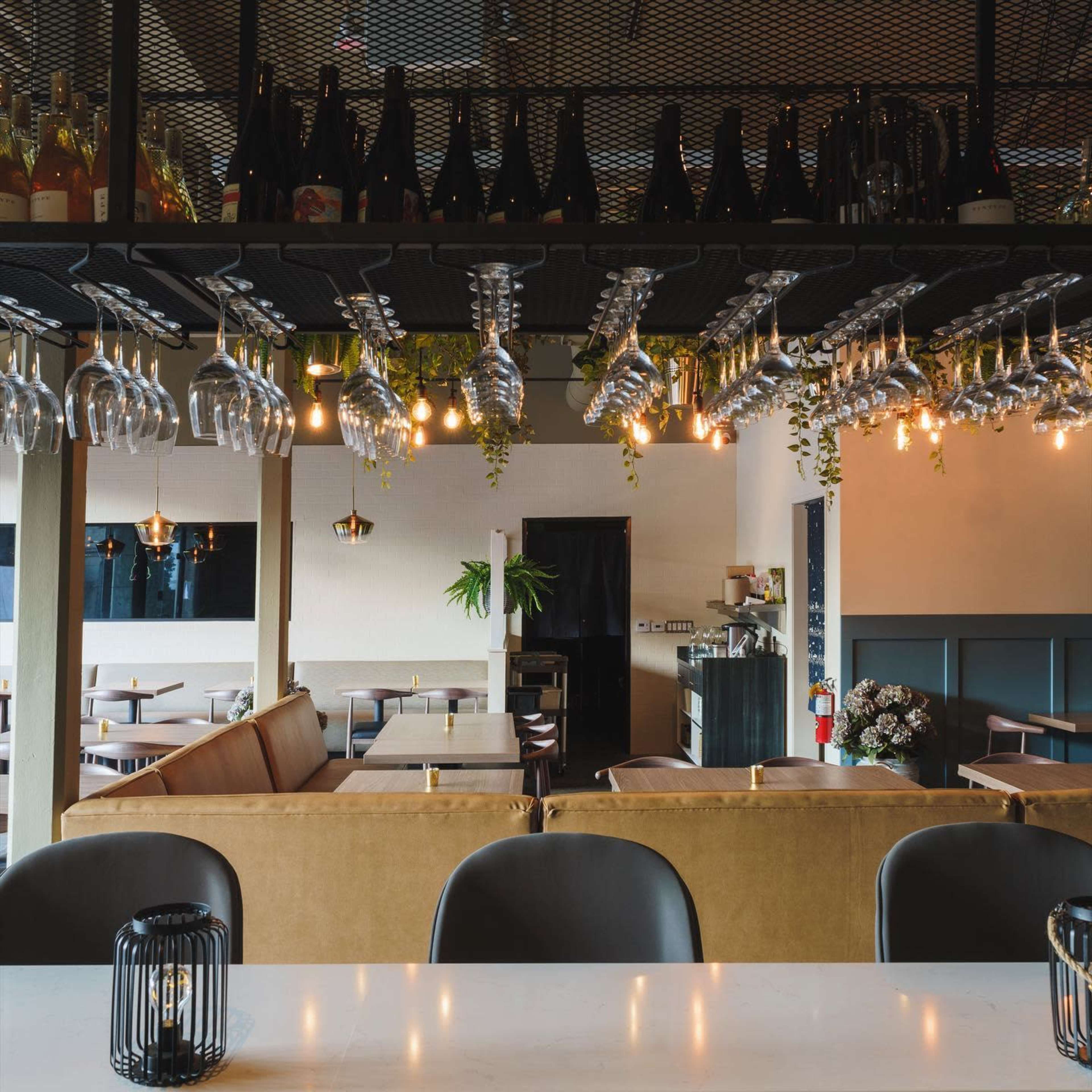 A modern restaurant interior with a bar area featuring hanging wine glasses and bottles, surrounded by tables and chairs.