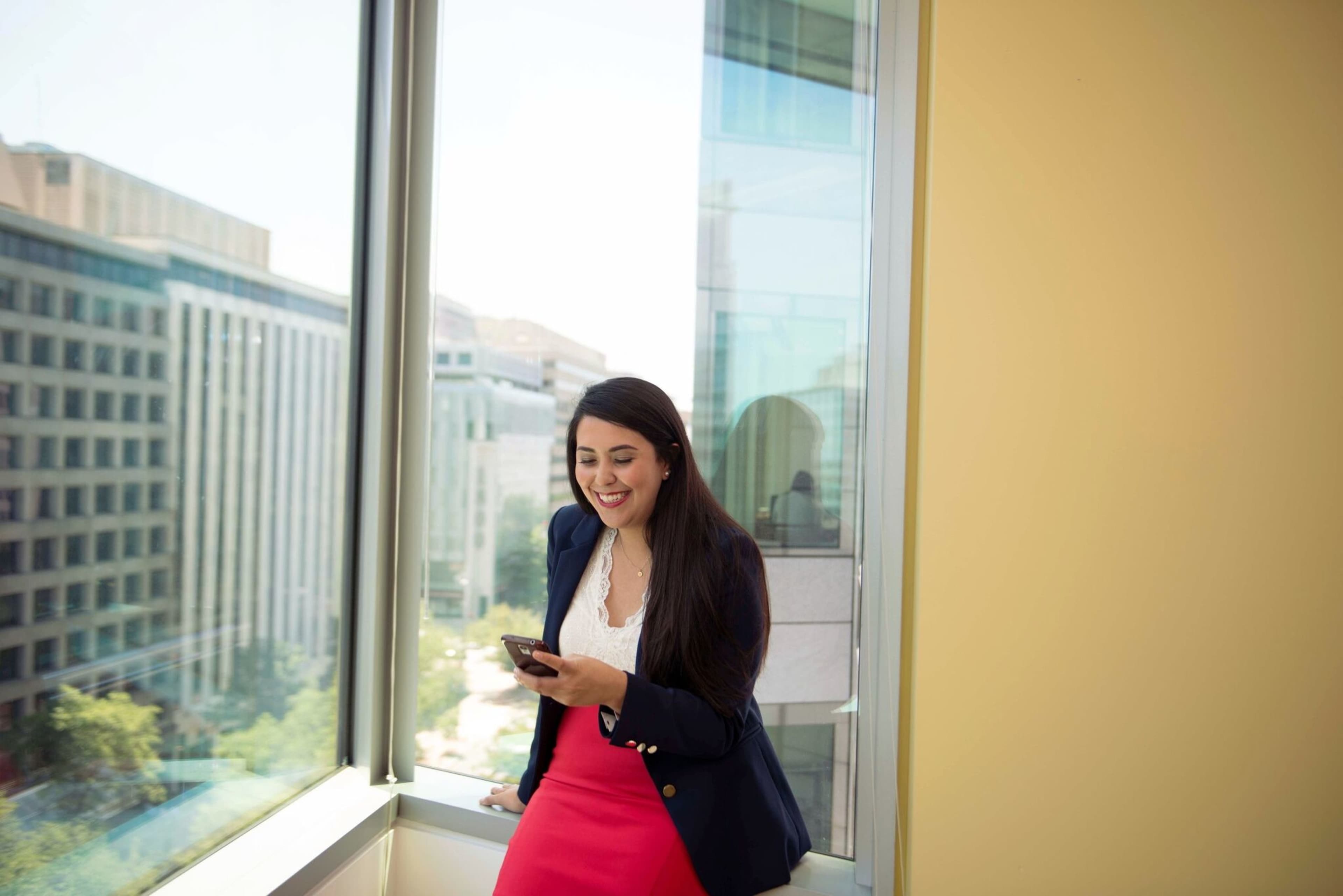 A woman in a blazer and pink skirt stands by a large window, looking at her phone with a cityscape in the background.