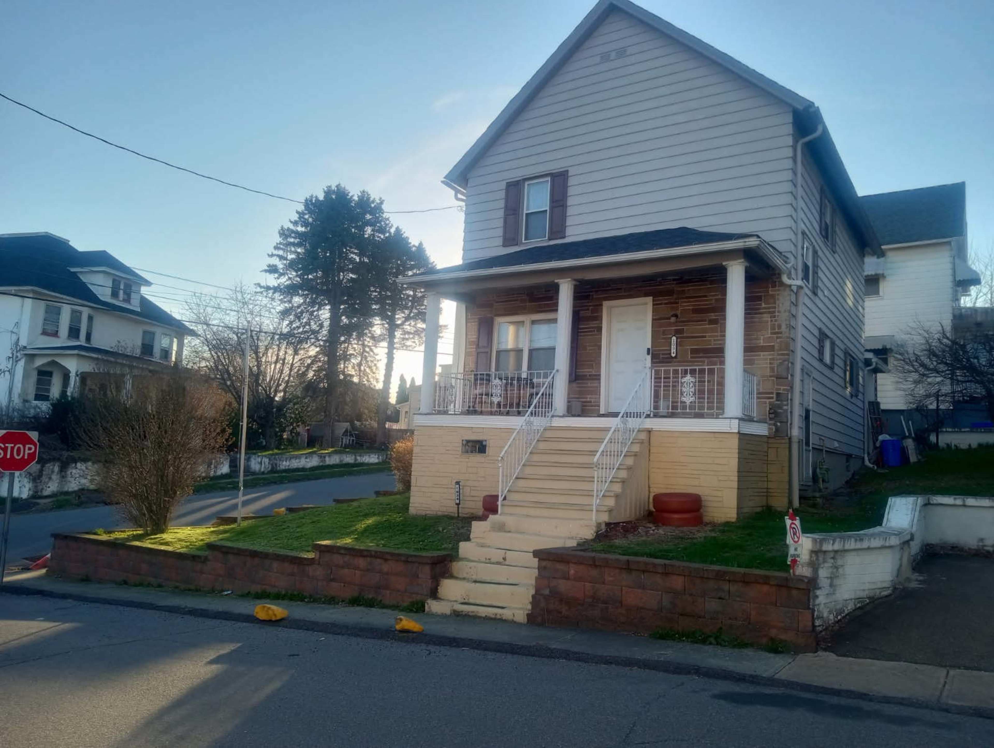 The image shows a two-story house with a front porch, steps leading to the entrance, and a small yard, situated on a residential street.