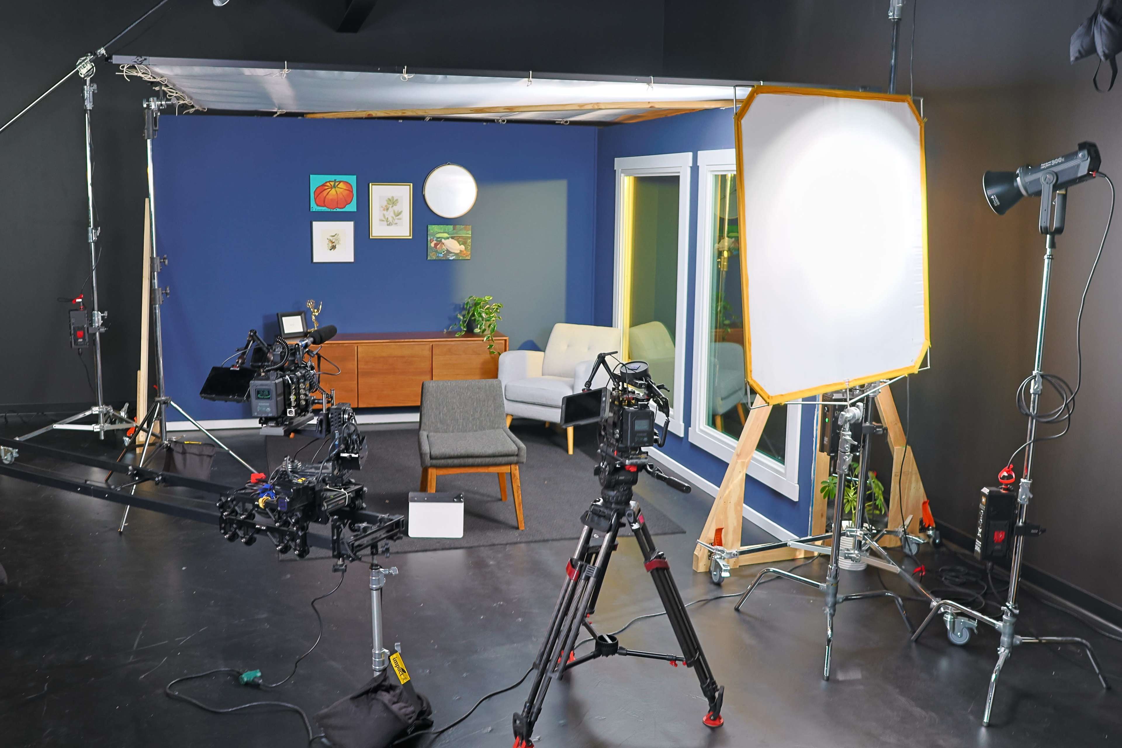 A film set features multiple cameras and lighting equipment arranged around a furnished room with a couch and decorative artwork on the walls.