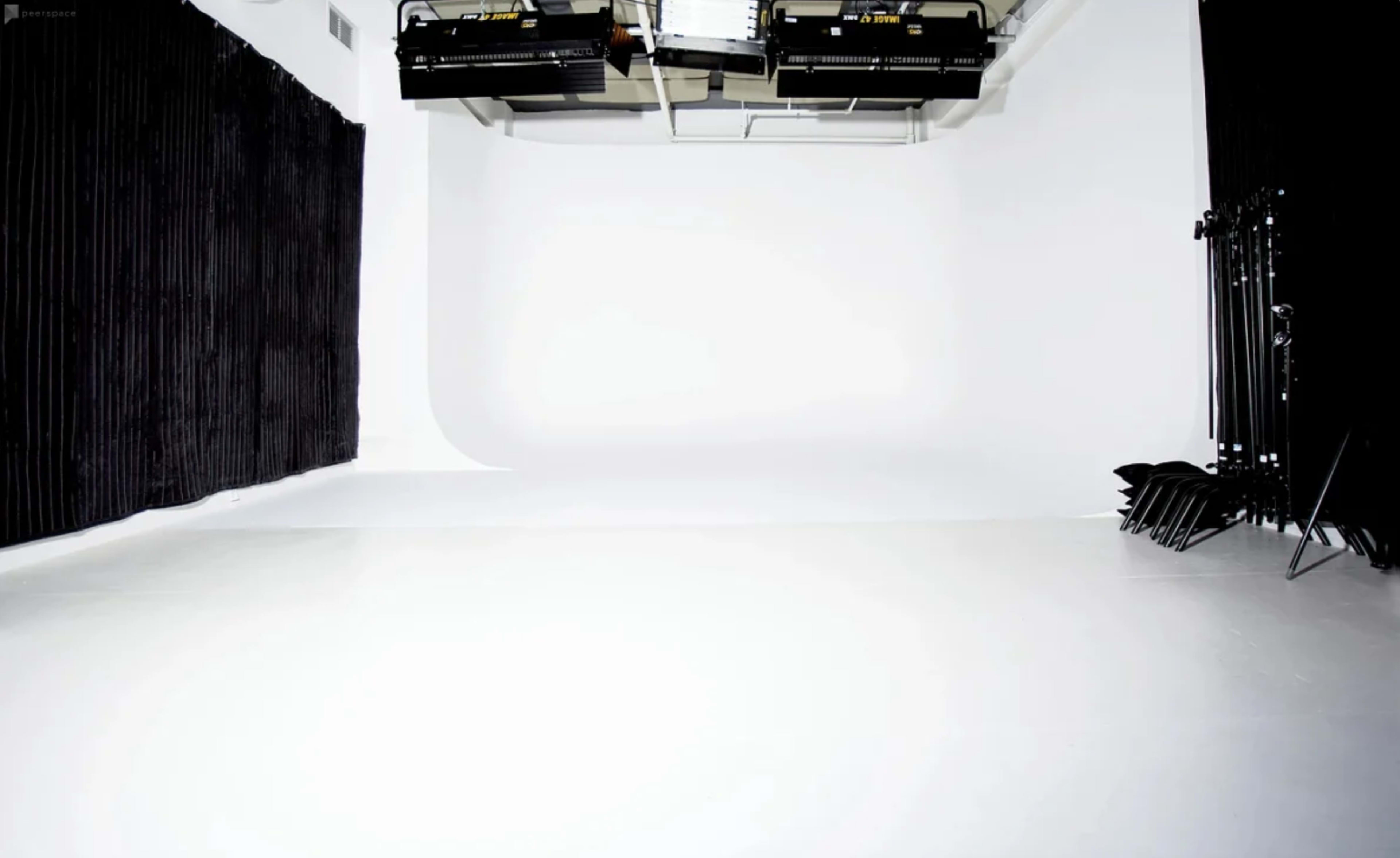 A photography studio with a white seamless backdrop, black curtains on one side, and studio lights mounted overhead.