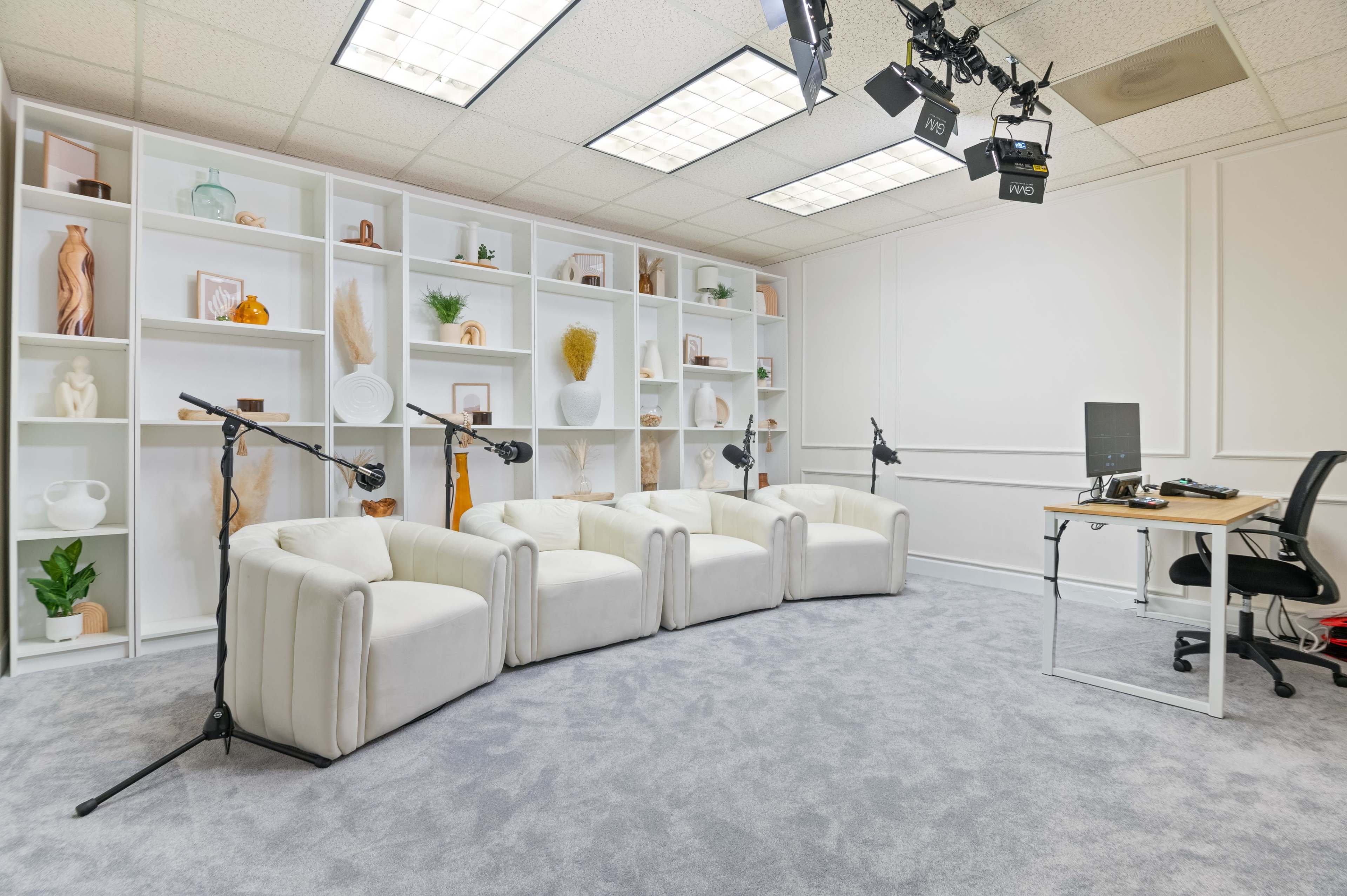 A modern interview set features three white chairs arranged in front of a bookshelf, with microphones on stands, a desk, and a chair in a well-lit room.
